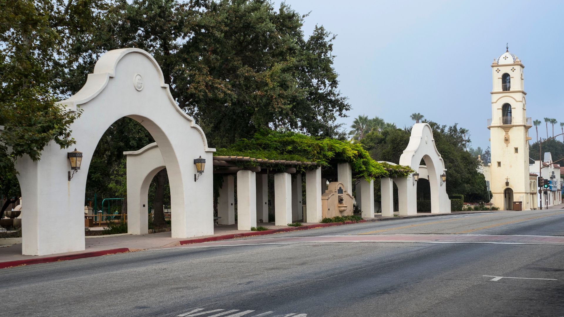 A wide-angle shot of a street in Ojai, California, featuring a grand white archway and a covered walkway (the Arcade) with a bell tower visible in the distance, all characteristic of Spanish Colonial Revival architecture.