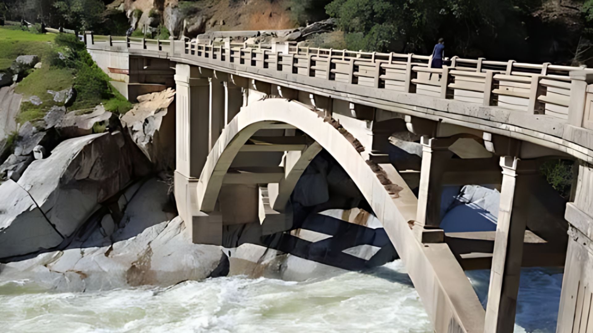 A concrete arch bridge, the Old Route 49 South Yuba River Bridge, spans over a swift-flowing river with large rocks visible in the water. Lush green trees and rocky cliffs line the riverbanks, and a person can be seen walking on the bridge in the distance.
