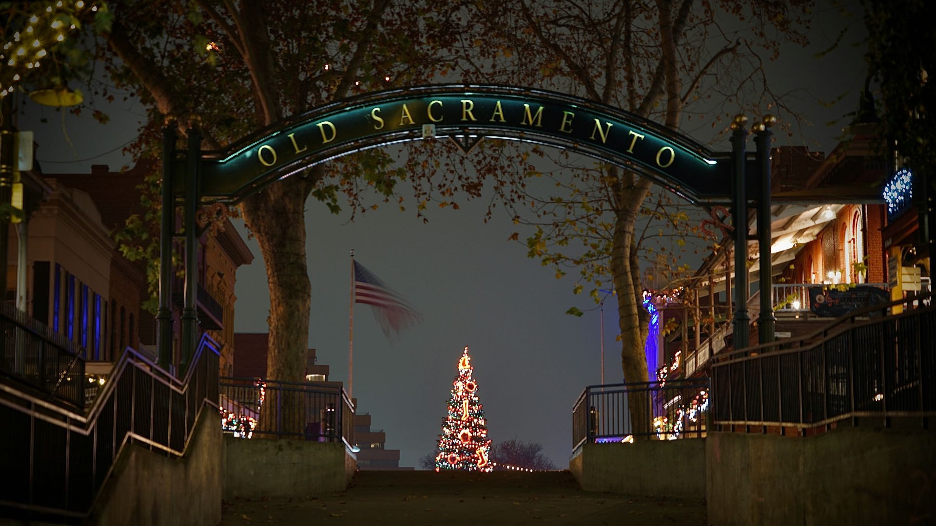 A nighttime view of the entrance to Old Sacramento, featuring a large archway sign illuminated with "OLD SACRAMENTO." In the distance, a tall, lit Christmas tree stands prominently, with an American flag visible to its left. The scene is framed by dark trees on either side and leads up a set of stairs or a sloping path.