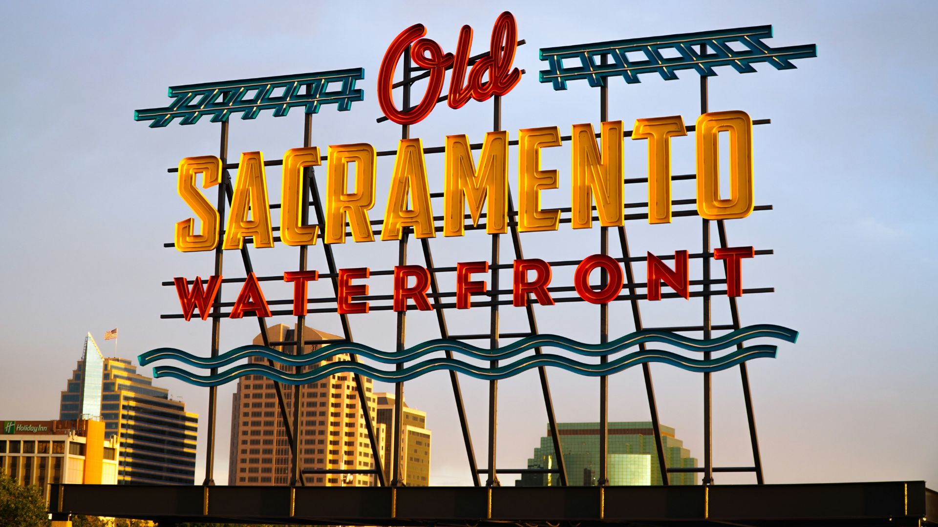 A large, illuminated sign in a vintage neon style reads "Old SACRAMENTO WATERFRONT" in red and gold letters, set against a clear sky with modern buildings visible in the background. The sign features wavy blue lines beneath the text, reminiscent of water, and is supported by a dark metal structure.