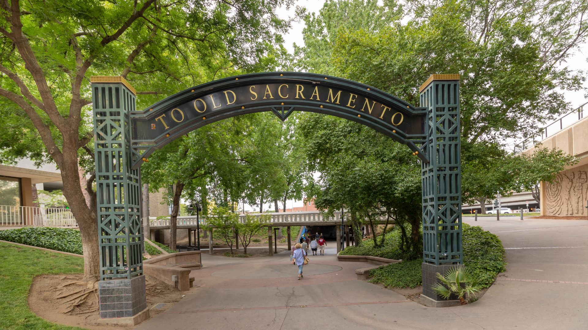 A large, dark archway with the words "TO OLD SACRAMENTO" in gold lettering spans a paved pathway. The arch is supported by two tall, ornate pillars. Lush green trees and foliage surround the pathway, with a bridge structure visible in the background to the right. A person is visible walking on the path beneath the arch.