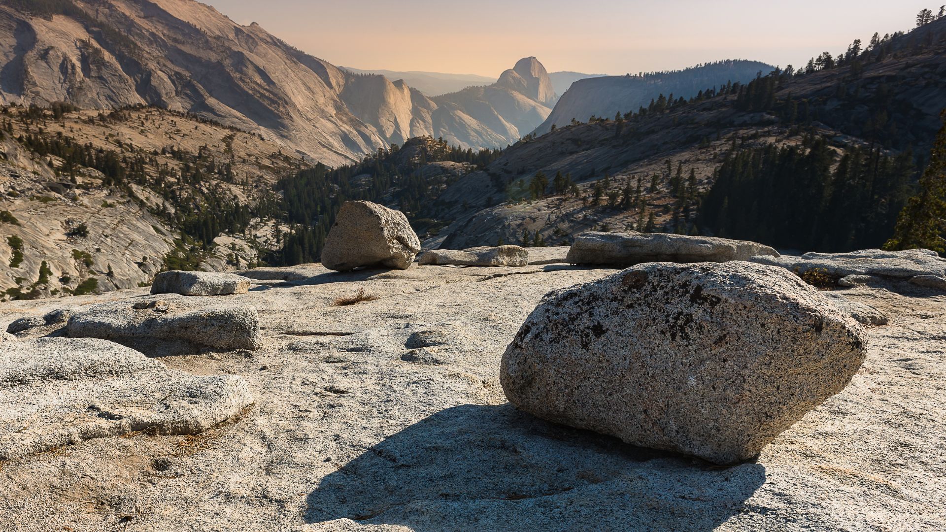 A wide-angle view of Olmsted Point in Yosemite National Park, featuring large glacial erratics on a granite slab in the foreground, and the iconic Half Dome visible in the distant background under a clear sky. 