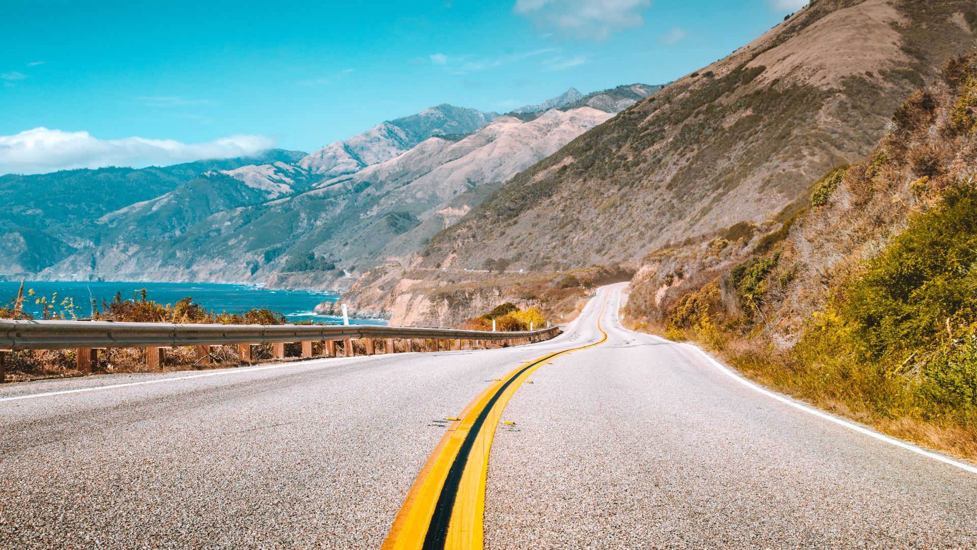 A scenic view of California's Pacific Coast Highway (Highway 1), winding along a dramatic coastline with mountains on the right and the Pacific Ocean visible on the left, under a clear blue sky.