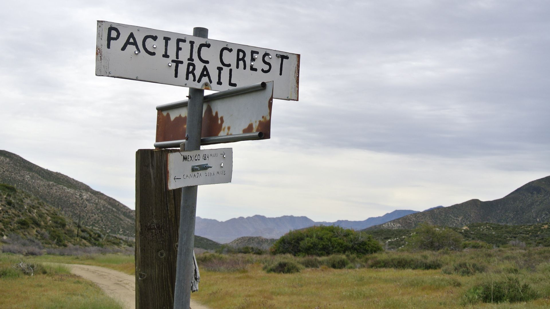 A weathered signpost in a rural, mountainous landscape points towards the "PACIFIC CREST TRAIL."
