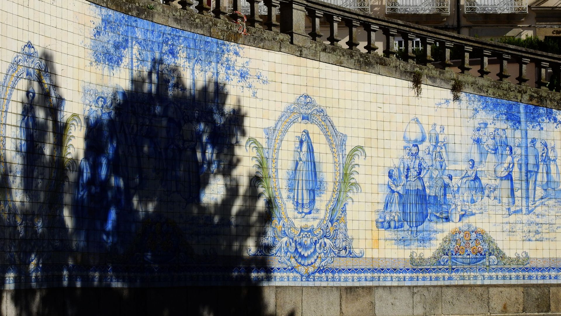 A vibrant blue and white azulejo tile mural on a wall in Viseu, Portugal, depicting scenes of rural life and religious figures, with a tree's shadow partially obscuring the left side.
