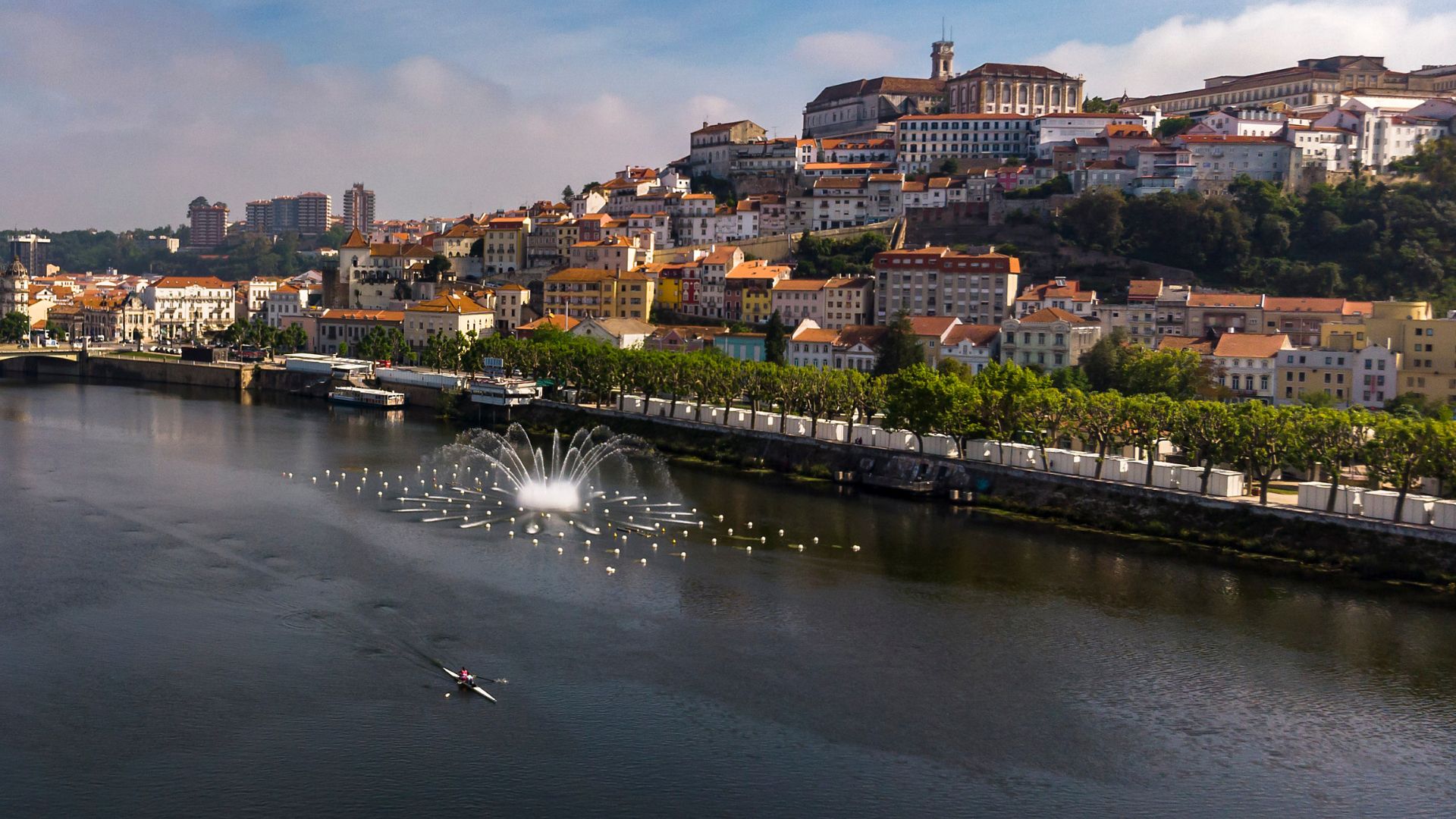 This image captures the scenic beauty of Coimbra, Portugal, with the historic city perched on a hill overlooking the Mondego River, where a fountain and a lone sculler add to the vibrant riverside scene of Parque Verde do Mondego.