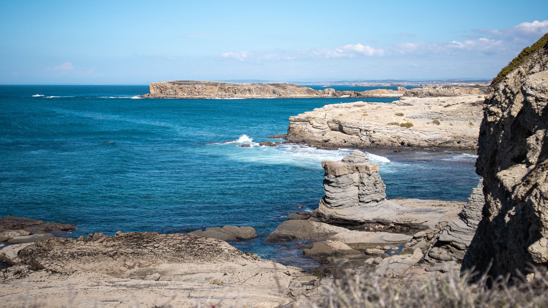 A vibrant coastal landscape in Peniche, Portugal, featuring a dramatic rocky shoreline, a prominent rock formation in the turquoise ocean, and distant cliffs under a clear blue sky. 