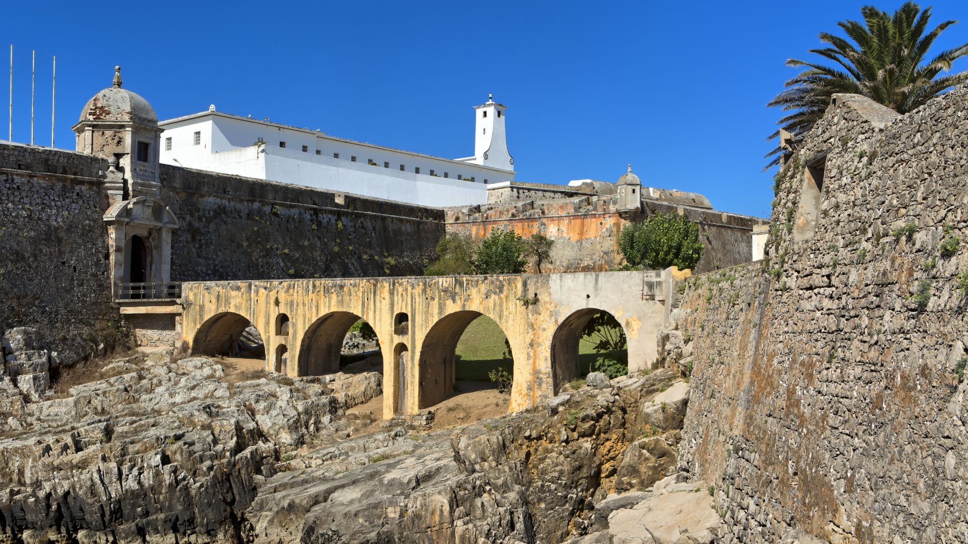 A panoramic view of the historic Peniche Fortress in Portugal, featuring a stone bridge with multiple arches spanning a rocky chasm, leading towards a large white building with a prominent tower and surrounding defensive walls under a clear blue sky.