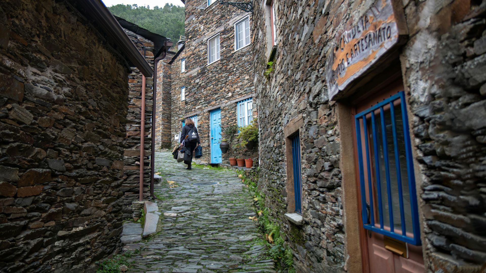 A narrow, cobblestone street winds through the historic village of Piódão in Portugal, lined with traditional schist stone houses and featuring a person walking away from the viewer.