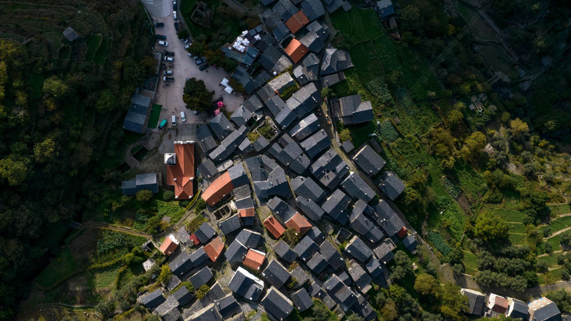 An aerial view of Piódão, a historic Portuguese village characterized by its distinctive schist houses with dark roofs clustered on a hillside, surrounded by lush green terraced slopes and winding roads.