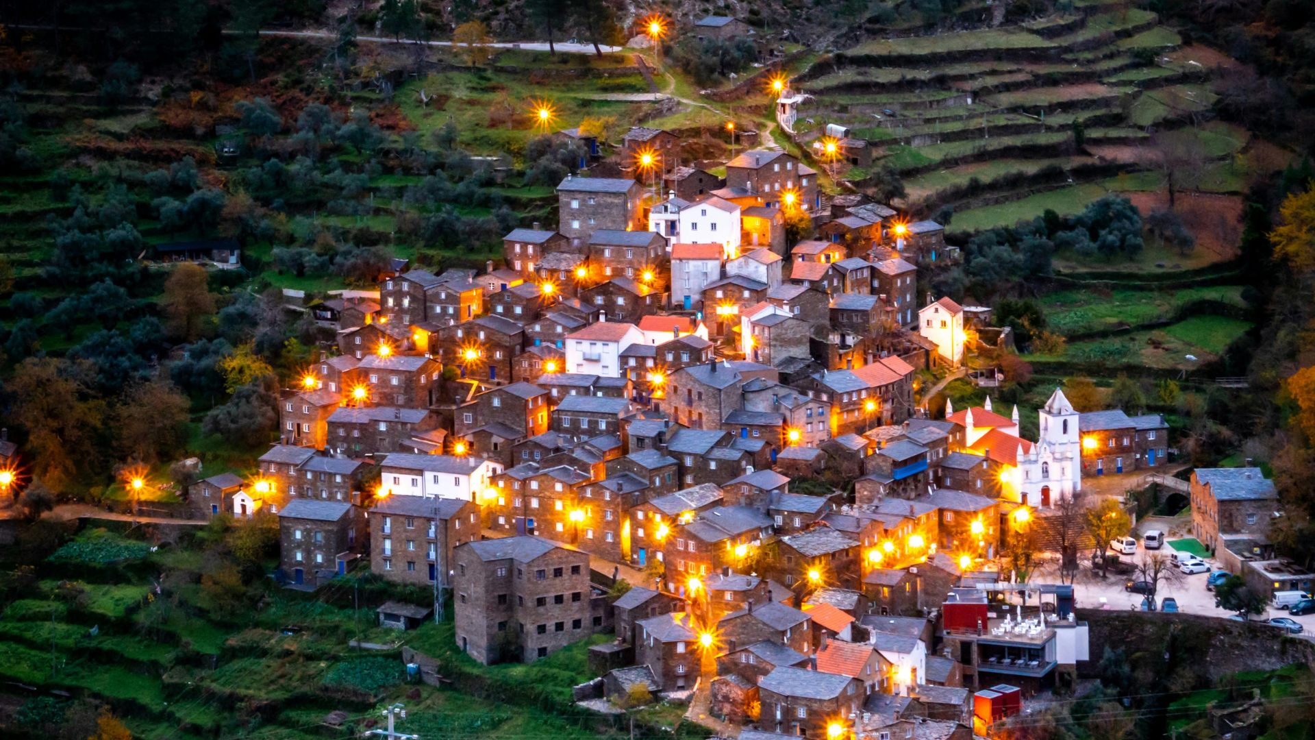 A nighttime aerial view of Piódão, a historic Portuguese village with schist houses and illuminated streets, nestled on a mountainside amidst terraced fields.