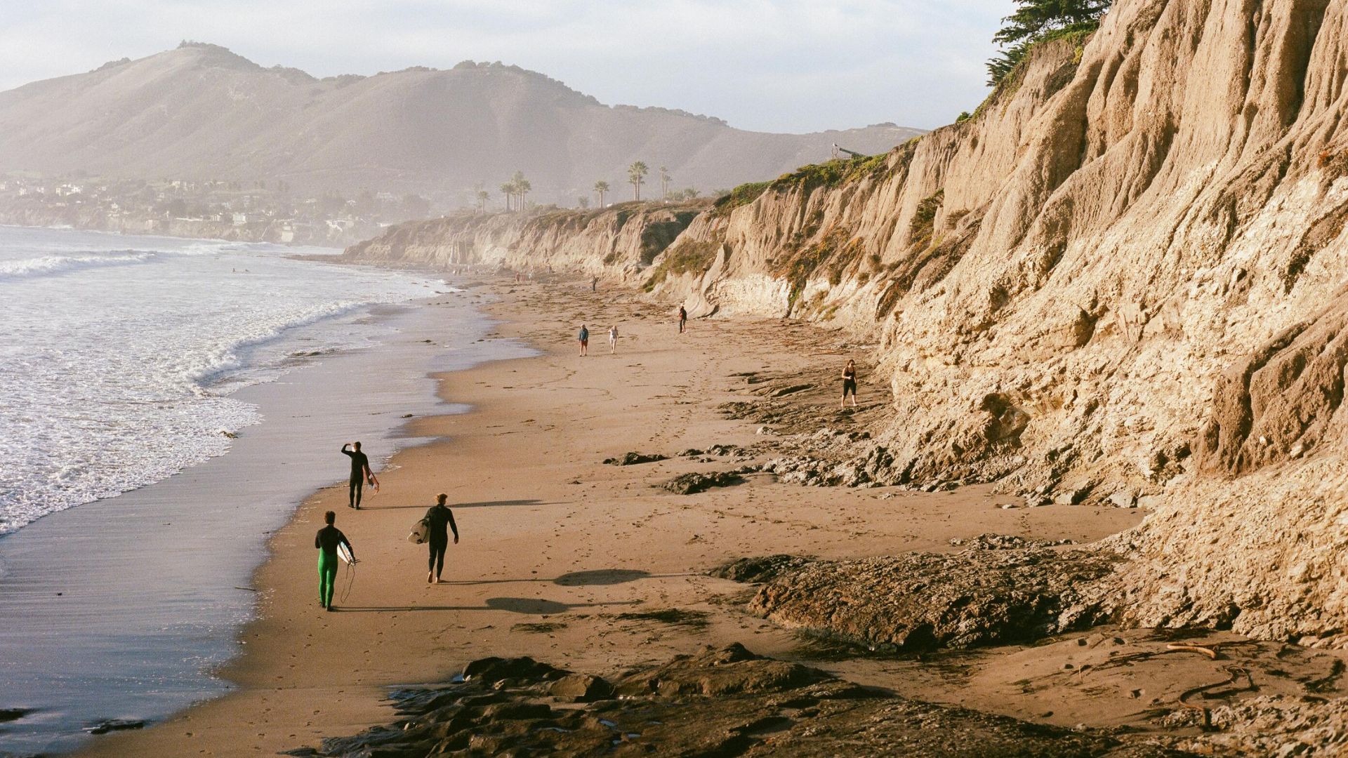A wide shot of Pismo Beach, California, showing a sandy shoreline with gentle waves on the left and dramatic, erosion-formed cliffs on the right. Several individuals, some carrying surfboards, are walking along the beach, with mountains visible in the hazy distance.