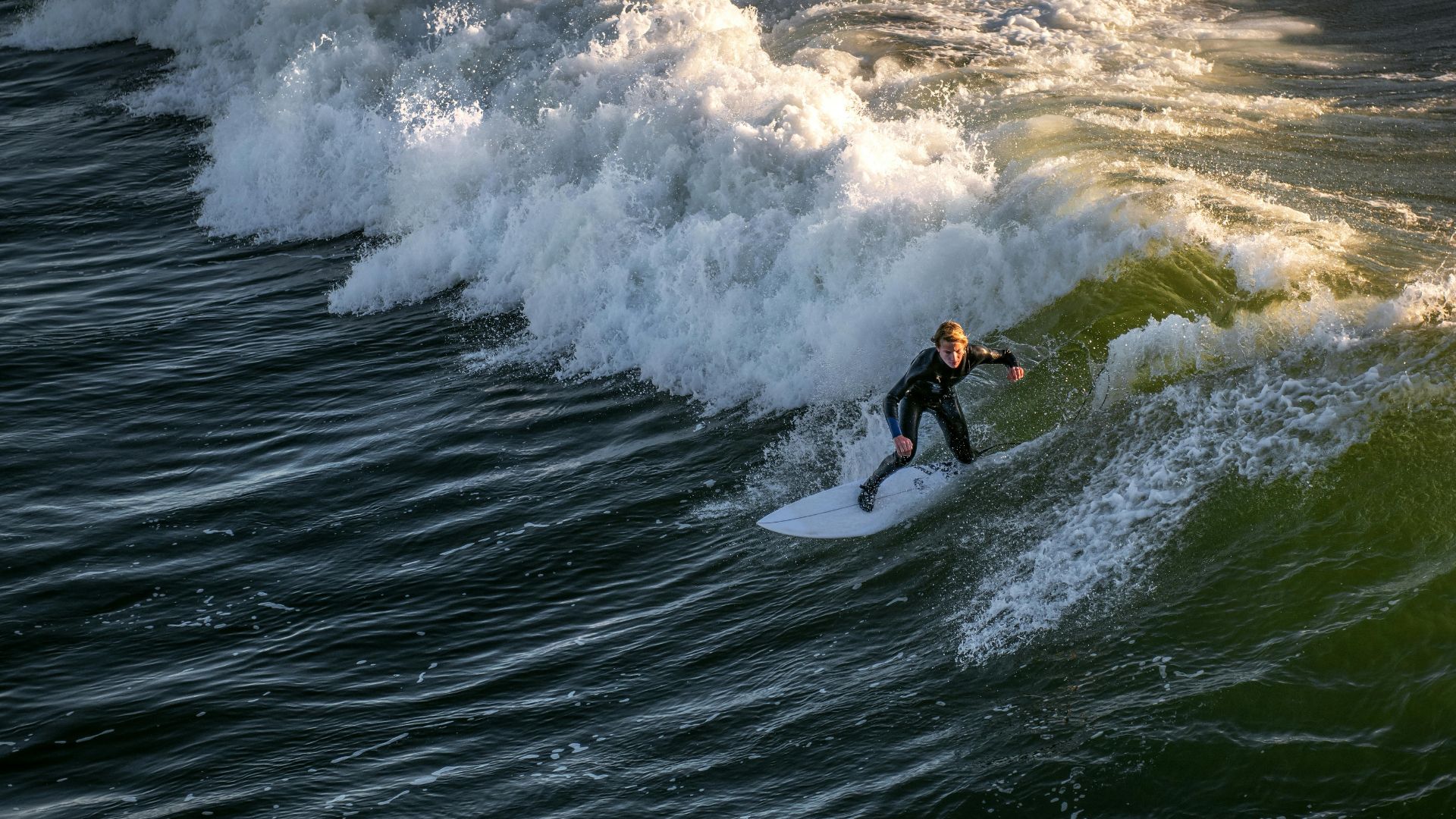 A surfer in a wetsuit skillfully rides a powerful wave on a surfboard, with the ocean water and white foam of the breaking wave visible around them, likely at a California beach known for surfing.