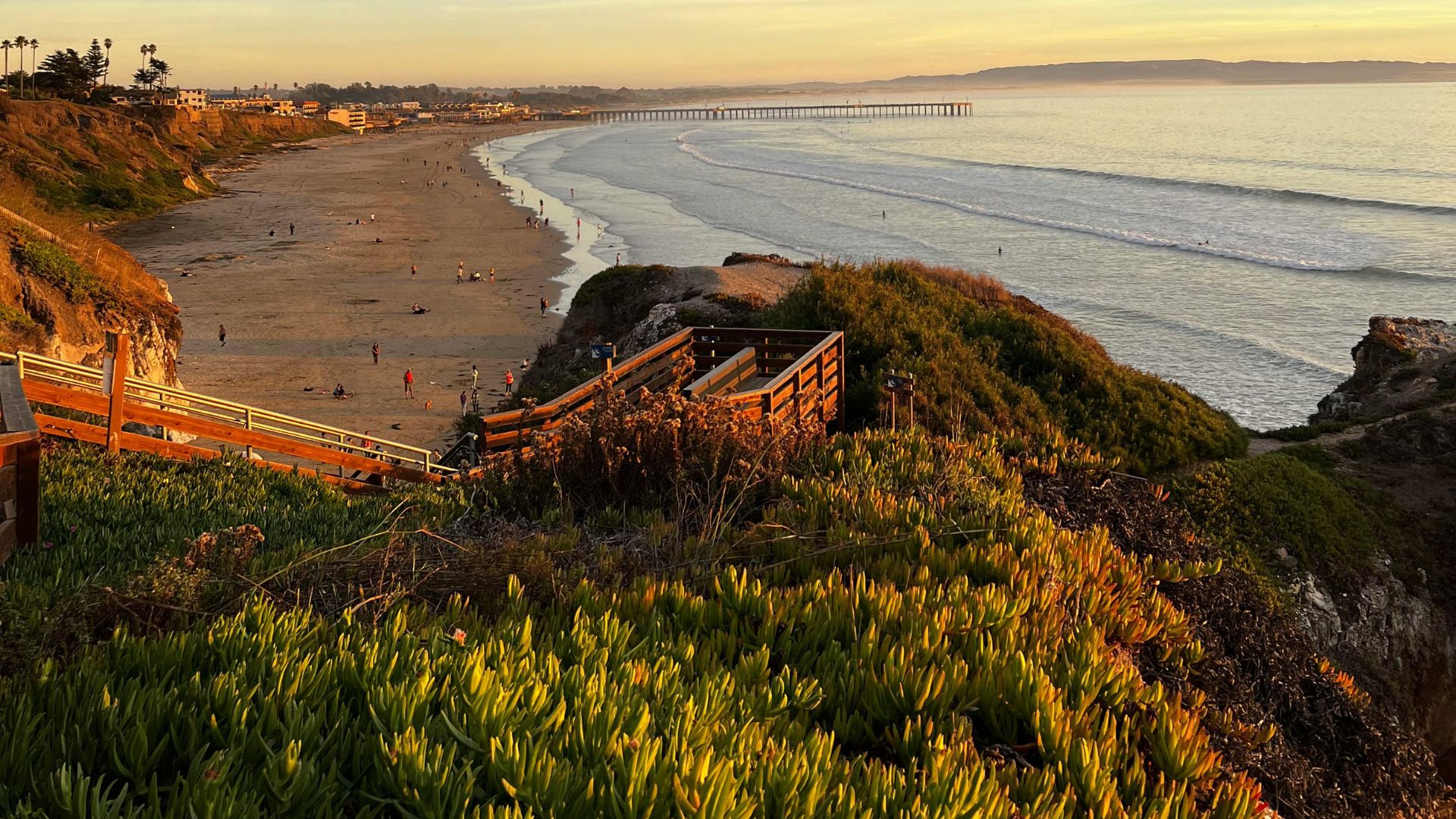 A vibrant sunset illuminates the sky over a wide sandy beach, with waves gently rolling towards the shore. In the foreground, lush green vegetation covers a cliffside, and a wooden staircase descends towards the beach. A long pier extends into the ocean in the distance under the colorful sky.