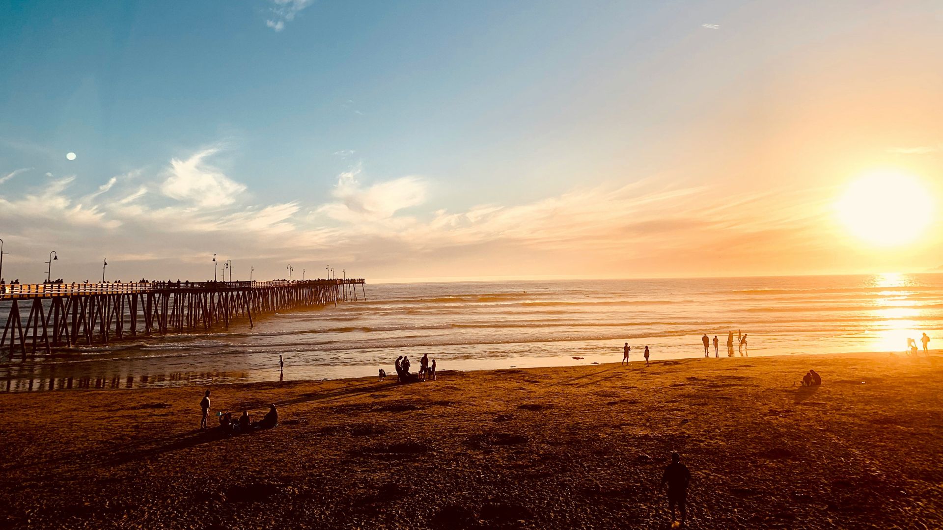 A sunset view of Pismo Beach, California, featuring a long pier extending into the ocean on the left, with waves gently rolling onto the shore where several people are scattered along the sand, enjoying the golden hour light.