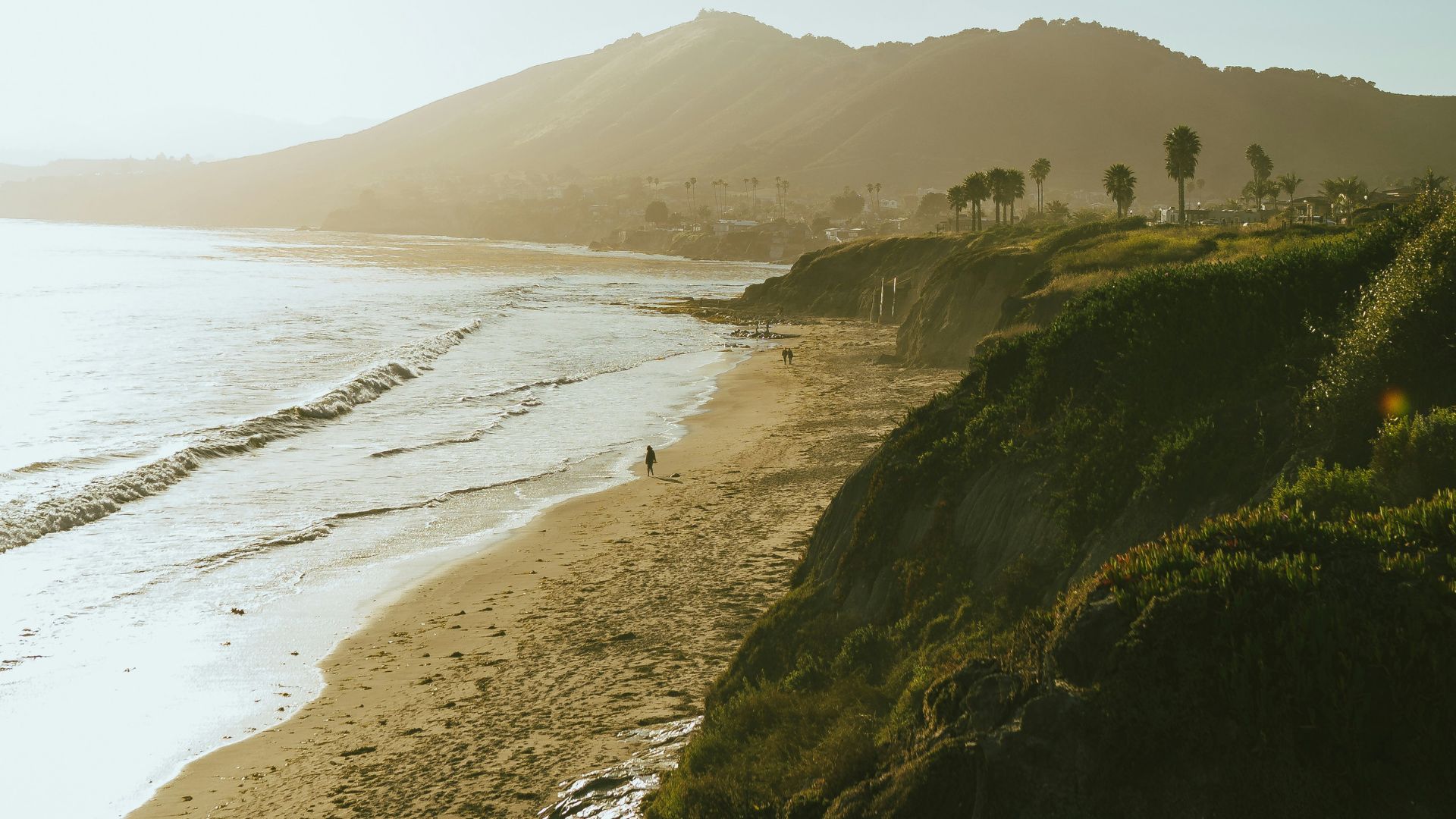 A sun-drenched, elevated view of a sandy beach and ocean waves crashing against the shore, with a person walking along the coastline in the distance. Lush green cliffs line the right side of the image, and a hazy mountain range rises in the background under a bright sky.