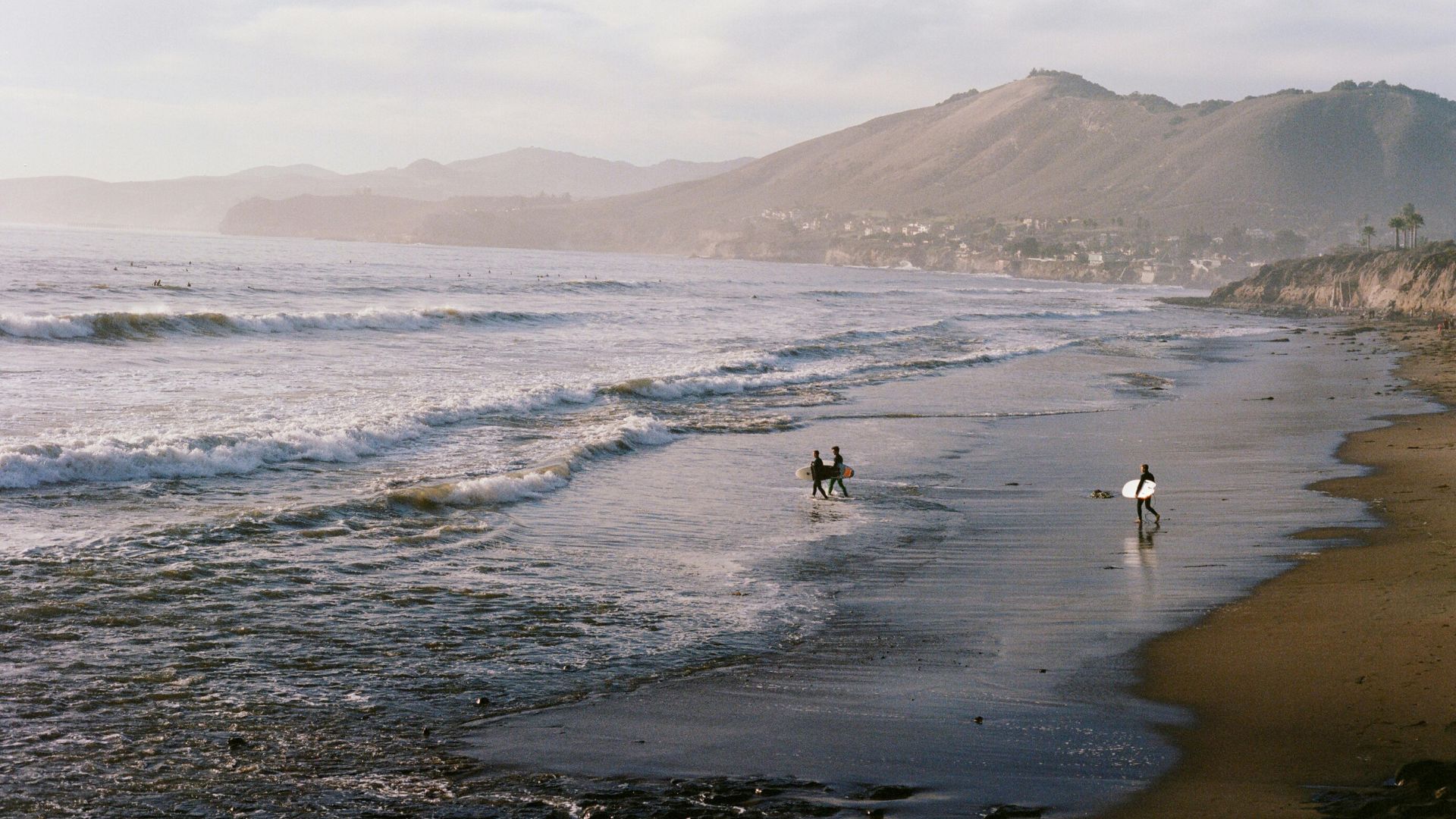 A wide shot of Pismo Beach in California, featuring waves breaking on the shore, two surfers walking near the water's edge, and mountains in the background under a hazy sky.
