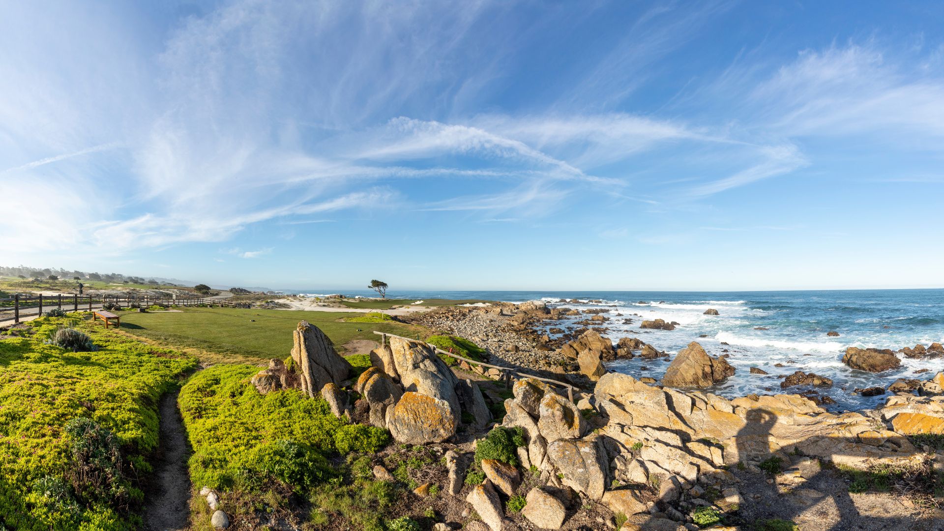A panoramic view of a rugged coastline with a rocky shore meeting the Pacific Ocean on a sunny day with wispy clouds in the sky. Lush green grass and a walking path are visible on the left, leading towards the ocean, with large granite rocks in the foreground and along the shoreline.