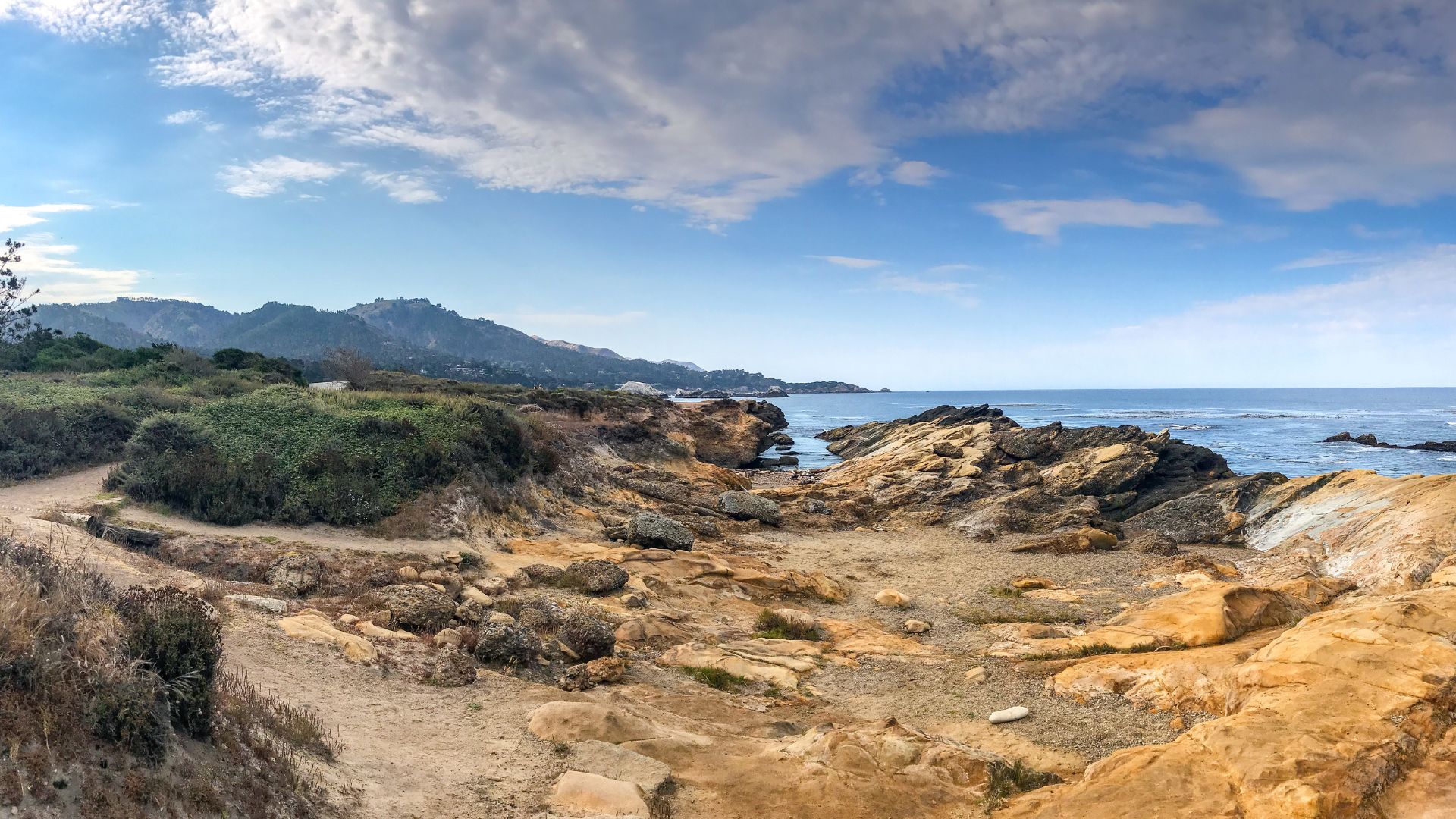 A panoramic view of the rugged, rocky coastline at Point Lobos State Natural Reserve, featuring a dirt path winding through low-lying green vegetation and brown, weathered rocks leading towards the Pacific Ocean under a partly cloudy blue sky.