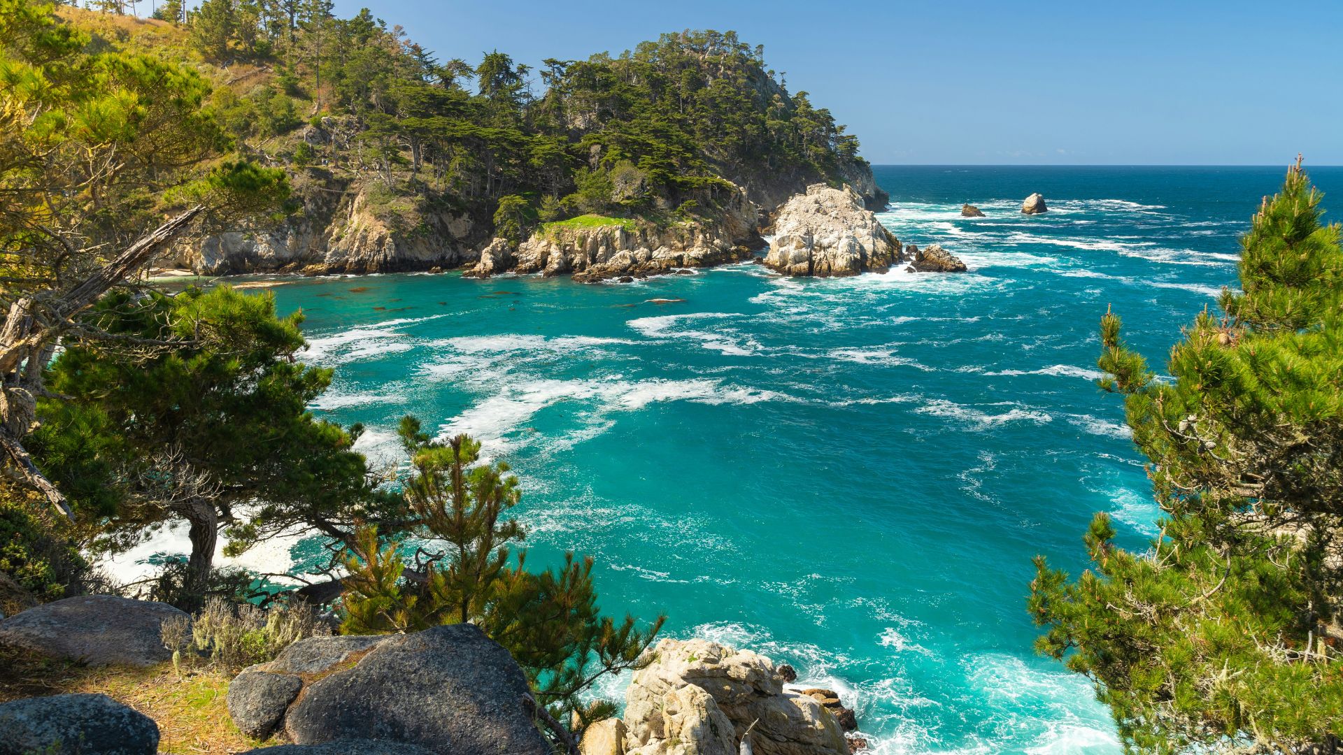 A scenic coastal landscape featuring turquoise ocean waters crashing against rocky shores and lush green cliffs covered in trees under a clear blue sky.