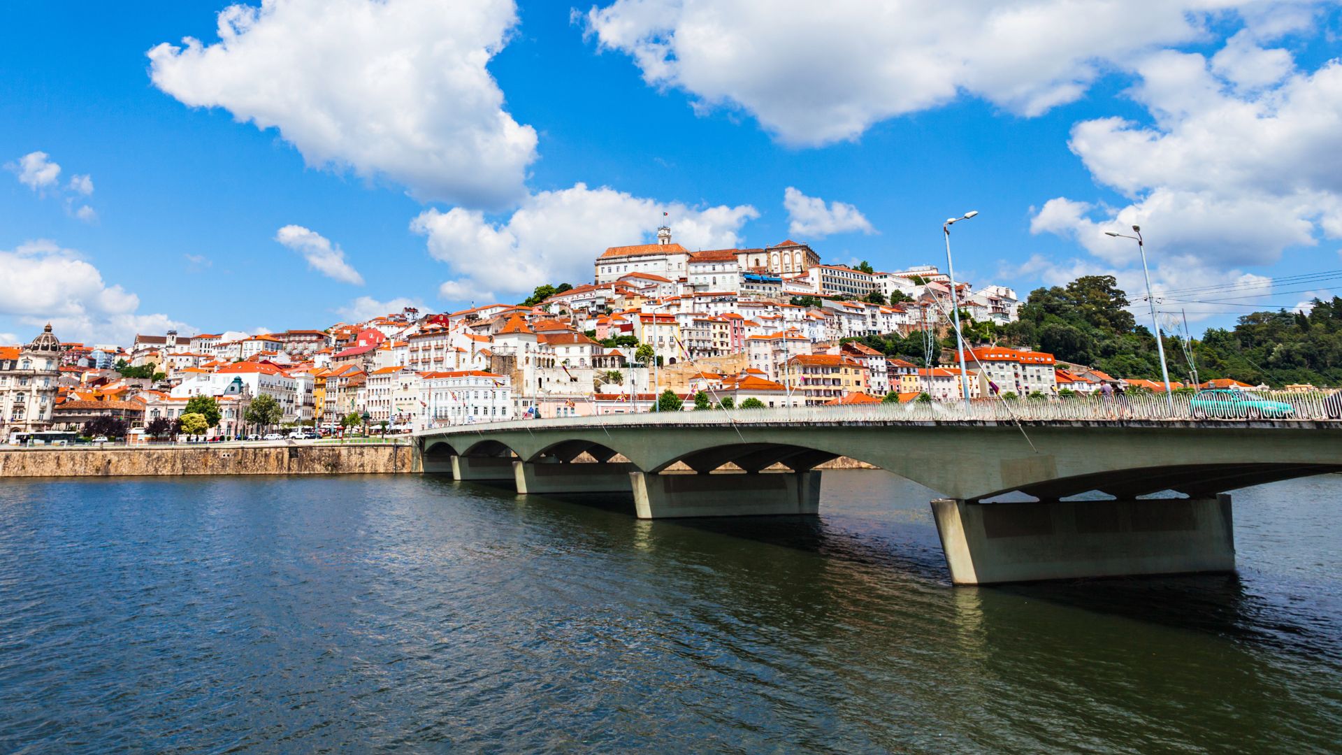 A panoramic view of Coimbra, Portugal, showcasing the historic city built on a hillside, with the Ponte de Santa Clara bridge crossing the wide Mondego River under a blue sky with white clouds.