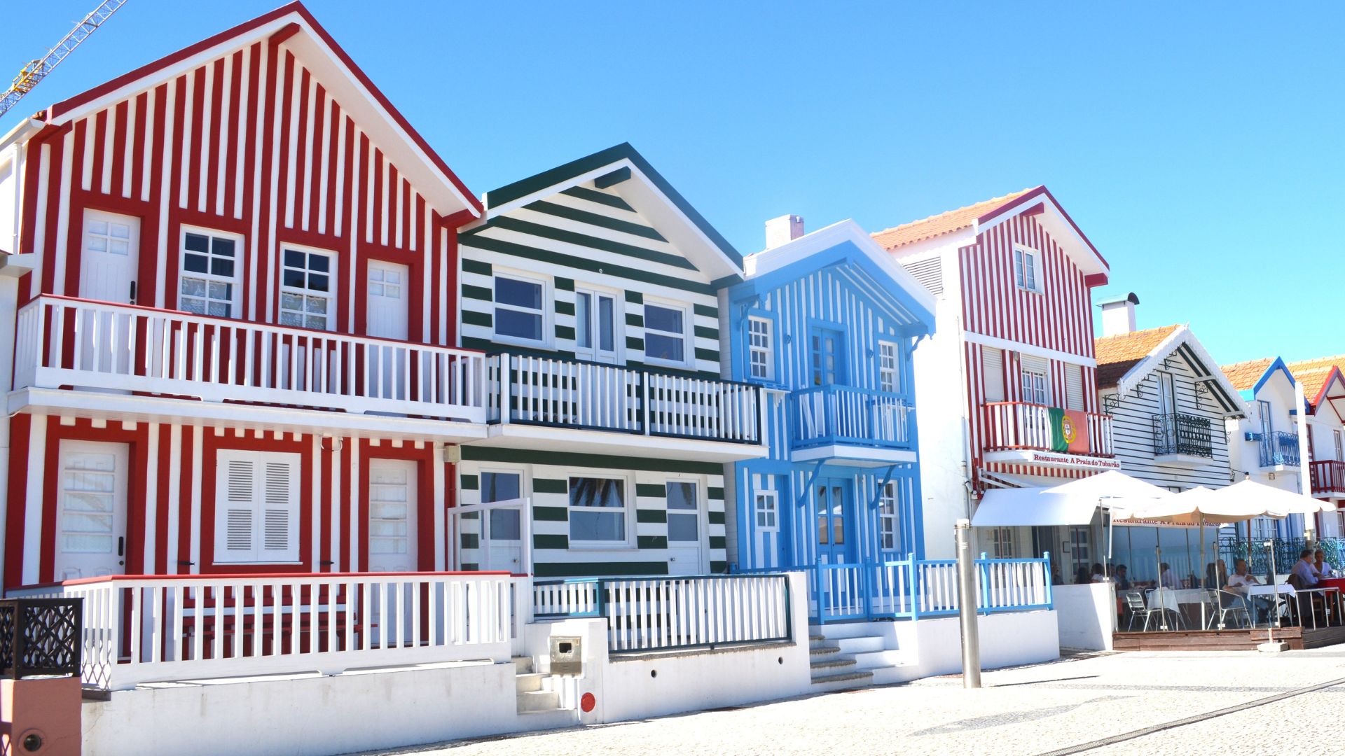 A row of colorful, vertically striped houses, known as "palheiros," in red and white, green and white, and blue and white, line a street under a clear blue sky in Costa Nova, Portugal. Some houses feature balconies with white railings, and a beach area with umbrellas and seating is visible on the right.