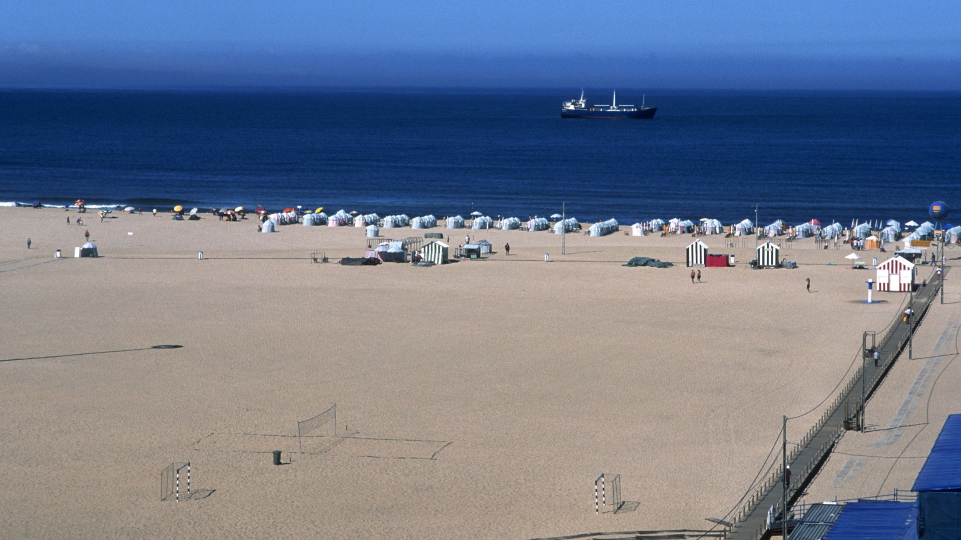 An aerial view of a wide, sandy beach with numerous white beach huts and a few distant ships on the calm blue ocean under a clear sky.