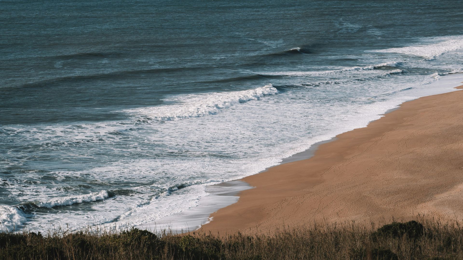 A wide-angle view of a sandy beach with large ocean waves breaking and receding, seen from a slightly elevated perspective.