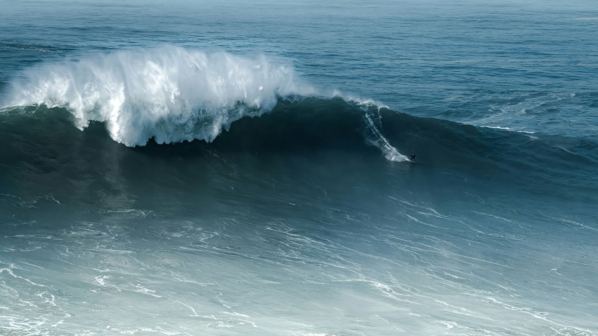 An aerial view of massive ocean waves crashing at Praia do Norte in Nazaré, Portugal, with several surfers visible in the water amidst the large swells.