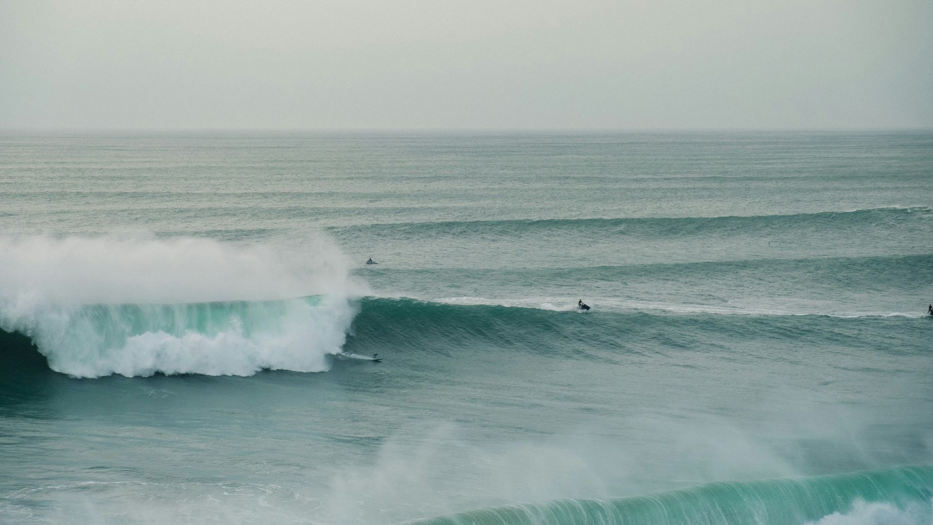 An aerial view of massive ocean waves crashing at Praia do Norte in Nazaré, Portugal, with several surfers visible in the water amidst the large swells.