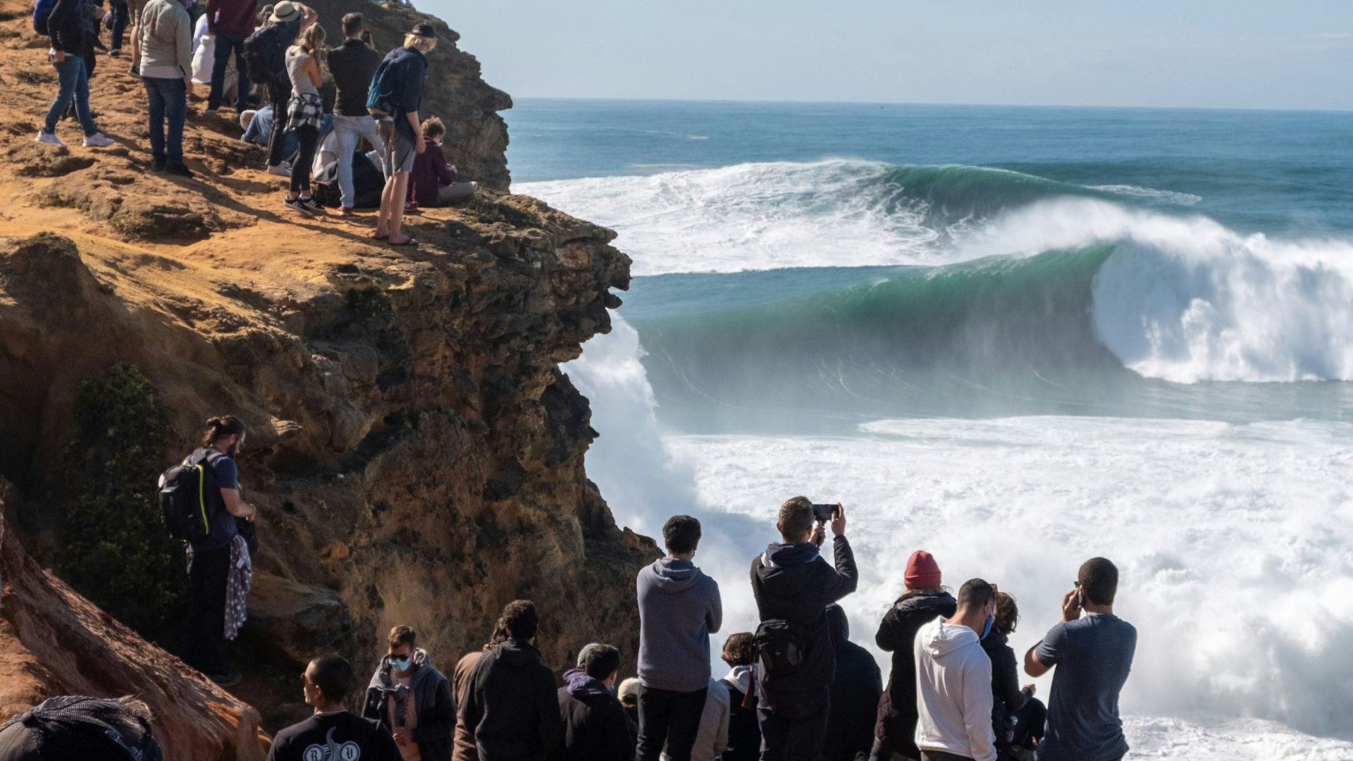  A crowd of people stands on a rocky cliff overlooking Praia do Norte in Nazaré, Portugal, watching massive waves break in the ocean below.