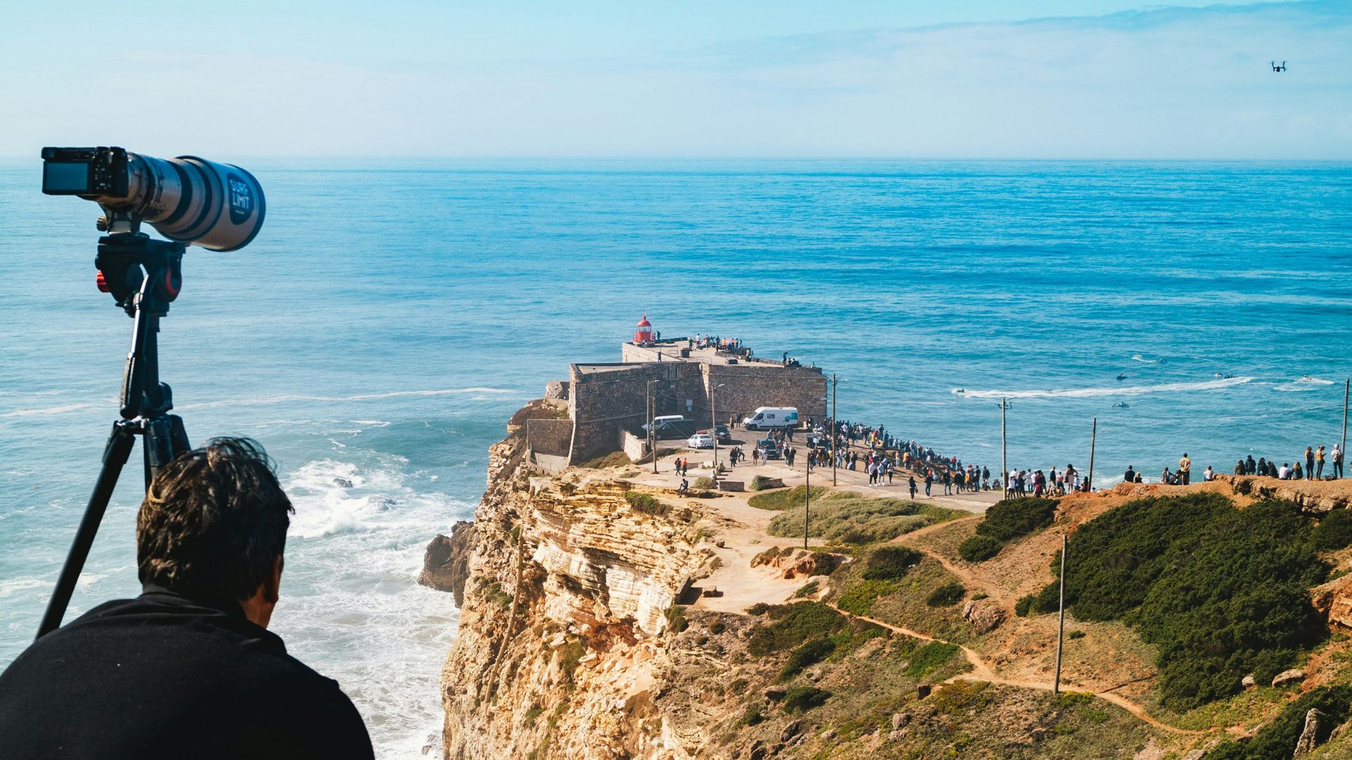  A person with a camera on a tripod overlooks a cliffside with a lighthouse and a crowd of people, with large waves crashing into the shore of Praia do Norte in Nazaré, Portugal.