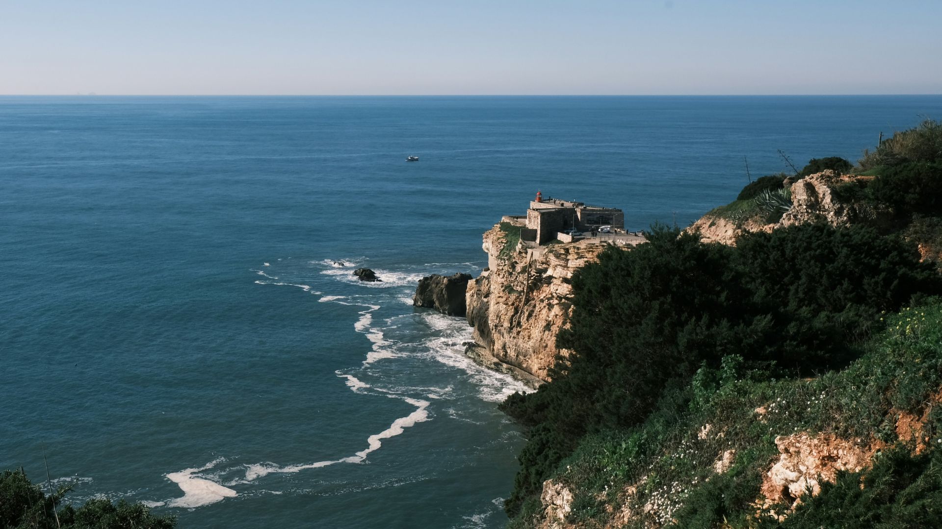 A scenic view of the Nazaré lighthouse perched on a cliff overlooking the vast blue Atlantic Ocean under a clear sky.