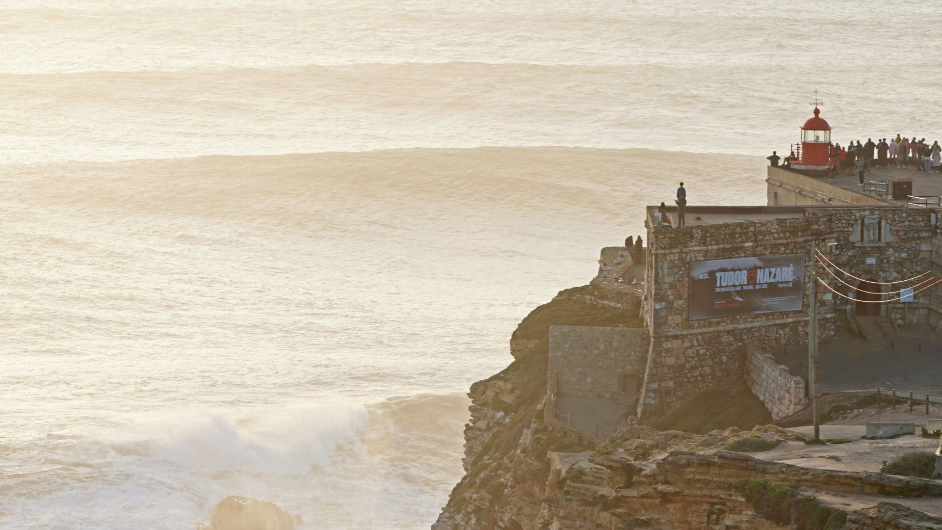 A scenic view of the Fort of São Miguel Arcanjo (Nazaré Lighthouse) atop a rugged cliff overlooking the ocean with large waves breaking at Praia do Norte in Nazaré, Portugal.
