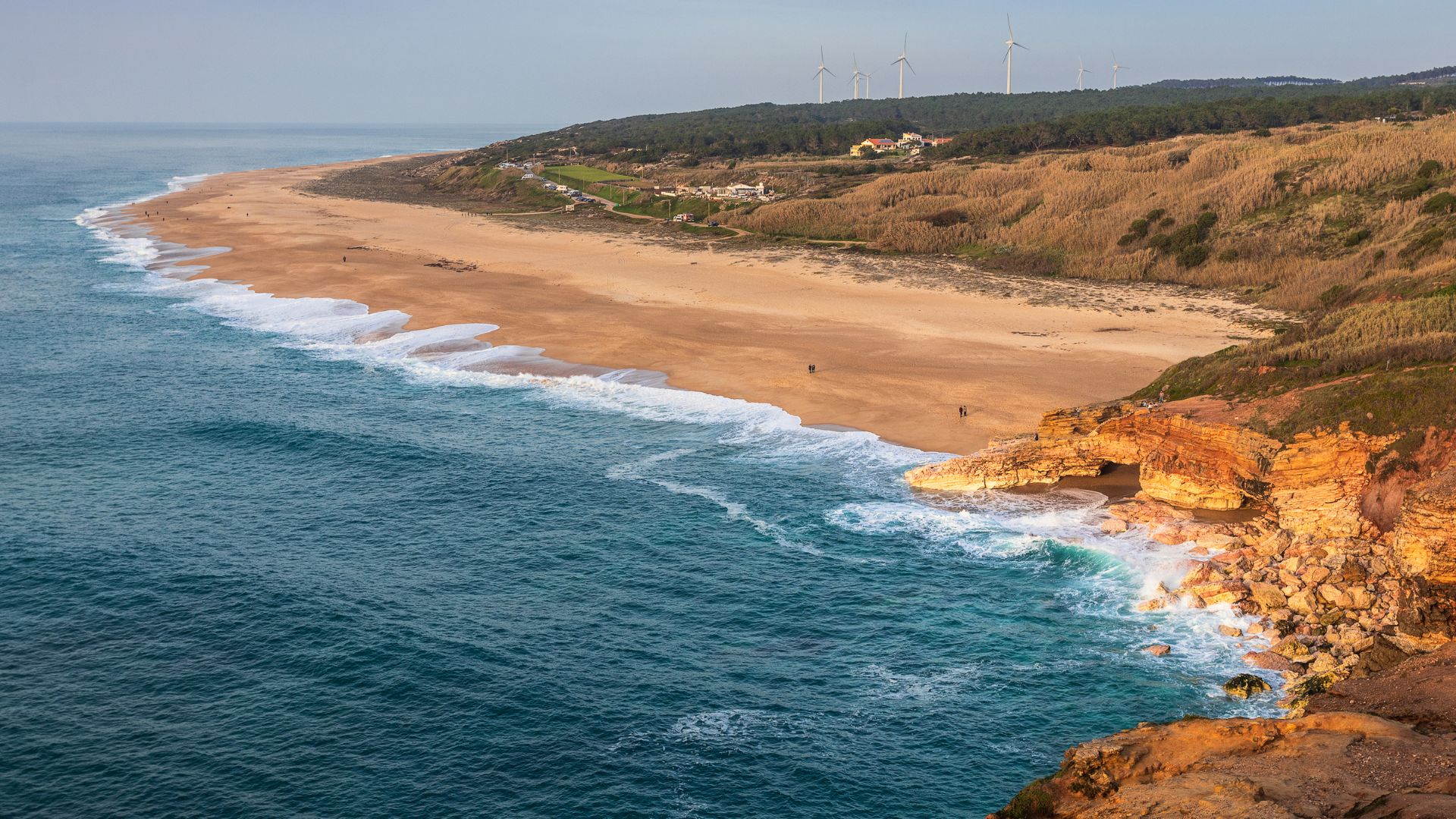 A wide-angle view of Praia do Norte (North Beach) in Nazaré, Portugal, showing a long stretch of golden sand, powerful waves crashing on the shore, and rugged cliffs with wind turbines visible in the distance. 