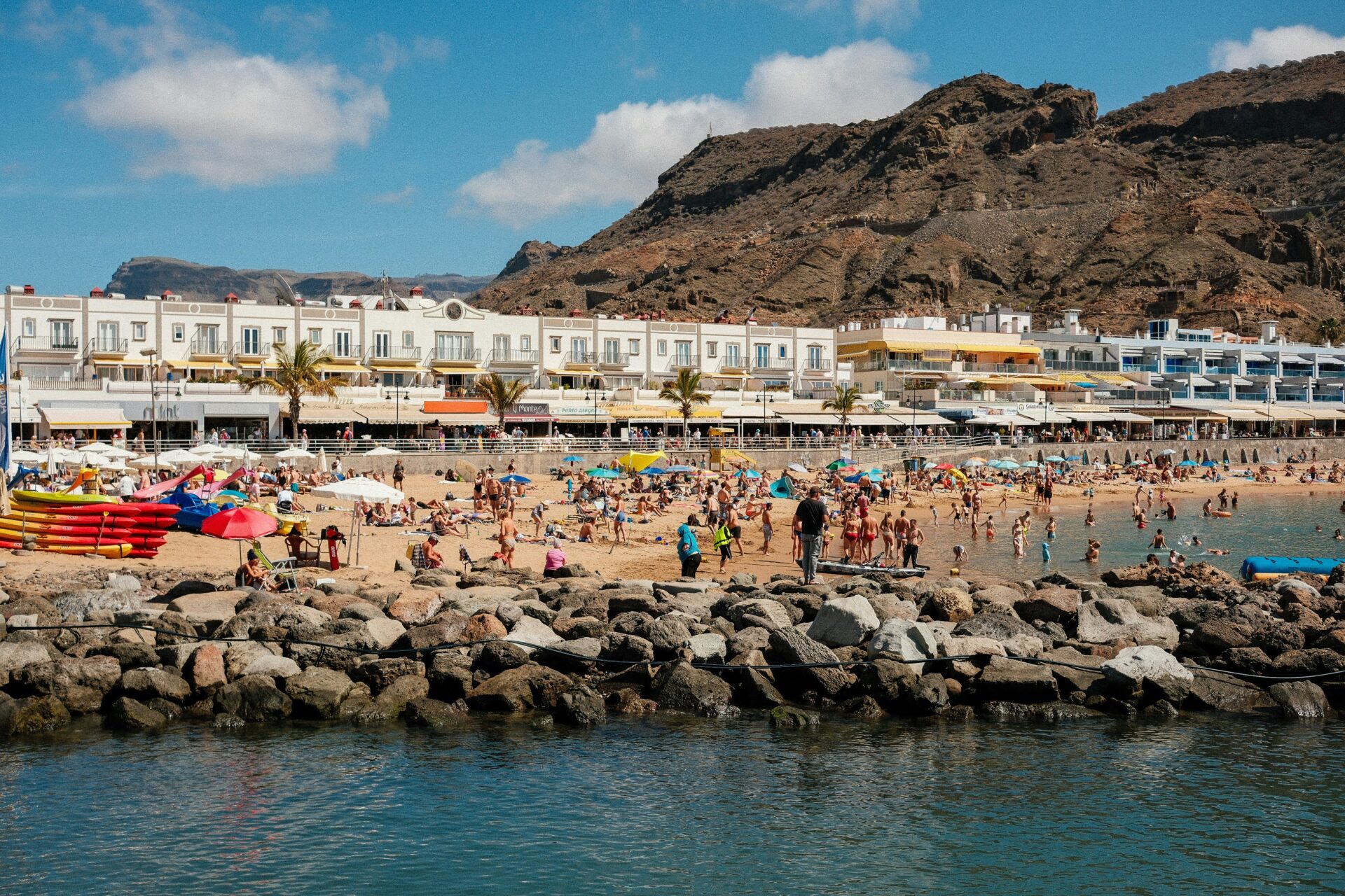 A stunning view of Puerto de Mogán, featuring whitewashed houses with colorful balconies and lush flowers, overlooking a serene beach with golden sand and crystal-clear waters.