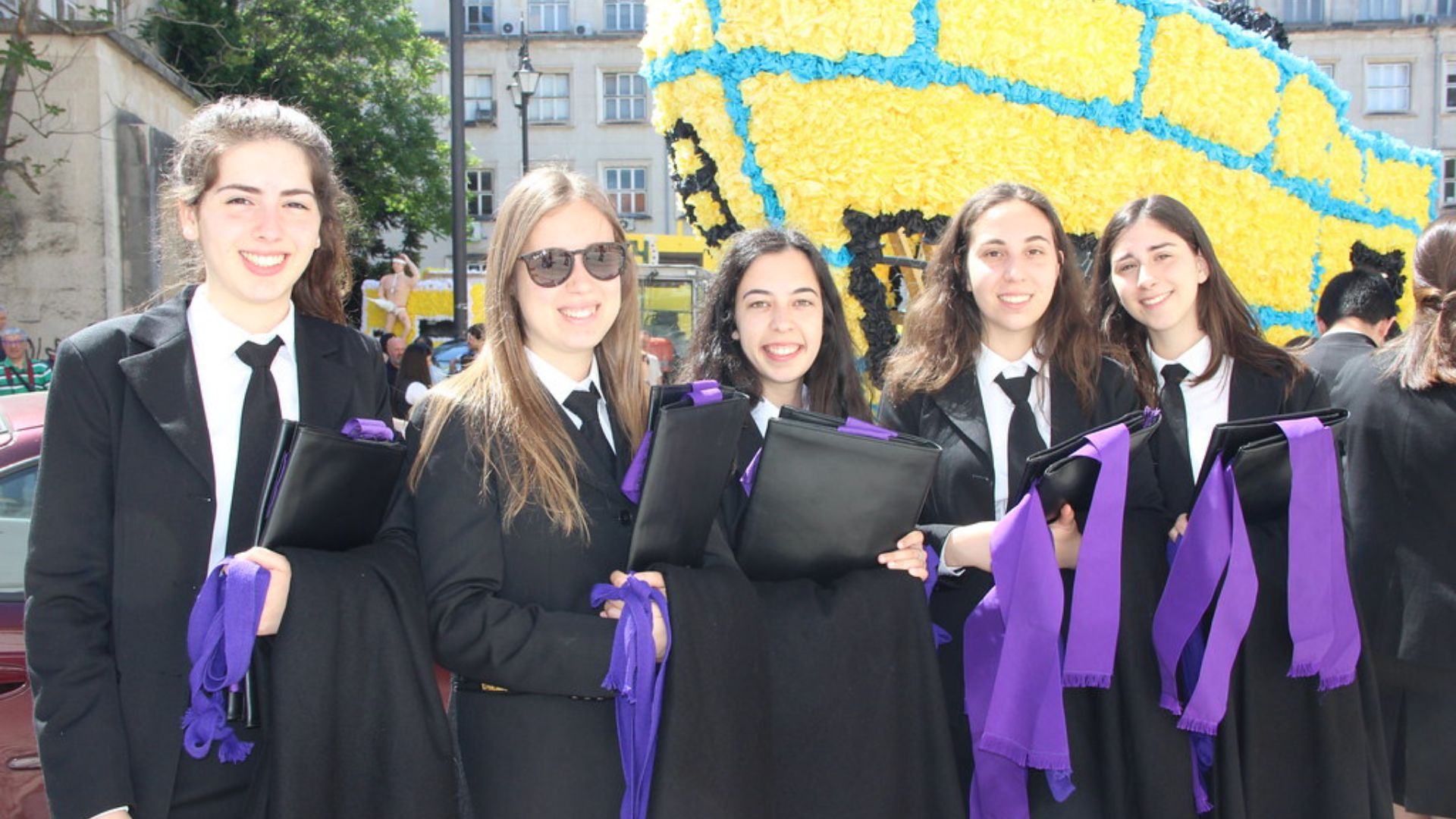 A group of smiling students in black academic robes and suits with purple sashes, holding folders, standing in front of a large, colorful float decorated with yellow and blue elements during the Queima das Fitas festival in Coimbra, Portugal.