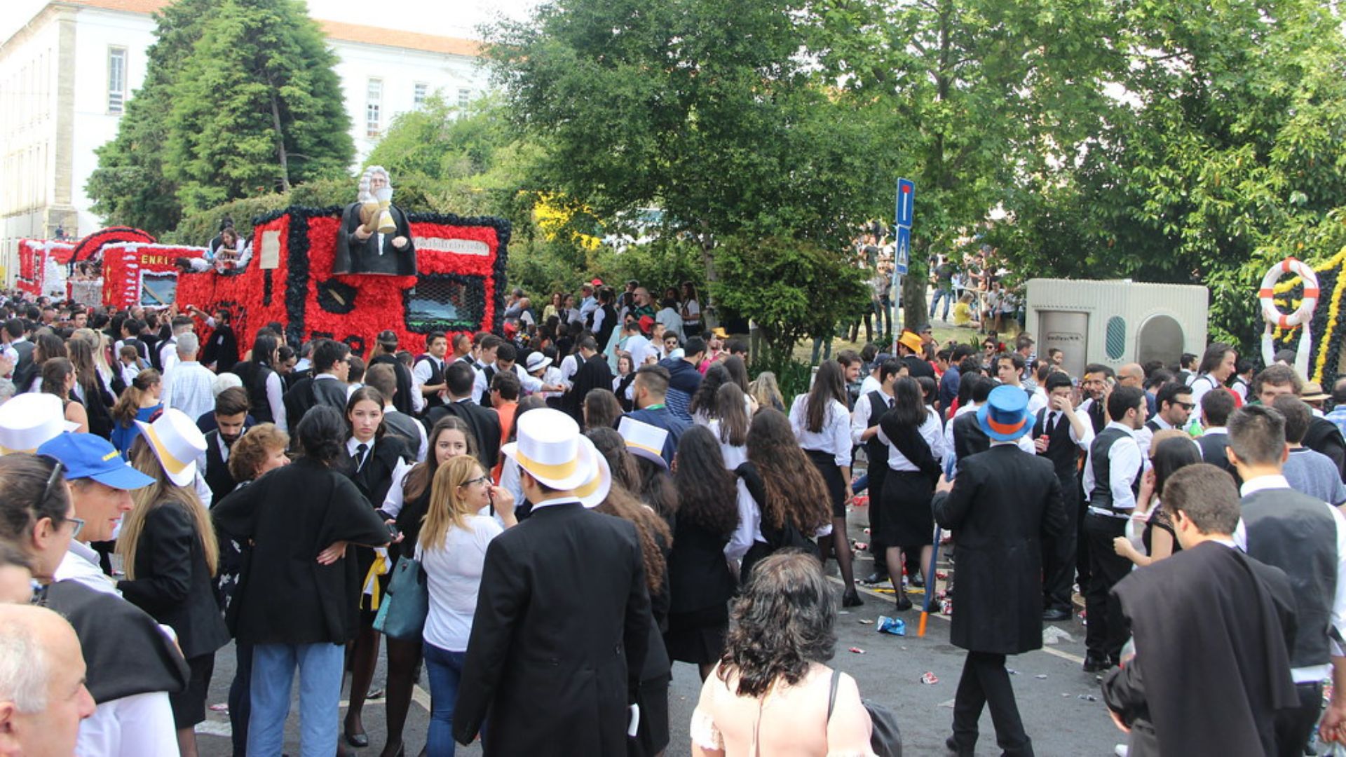 A large crowd of university students in black academic attire and some wearing hats, gathered outdoors during a daytime parade, with a large, elaborate red float topped with a figure in the background.