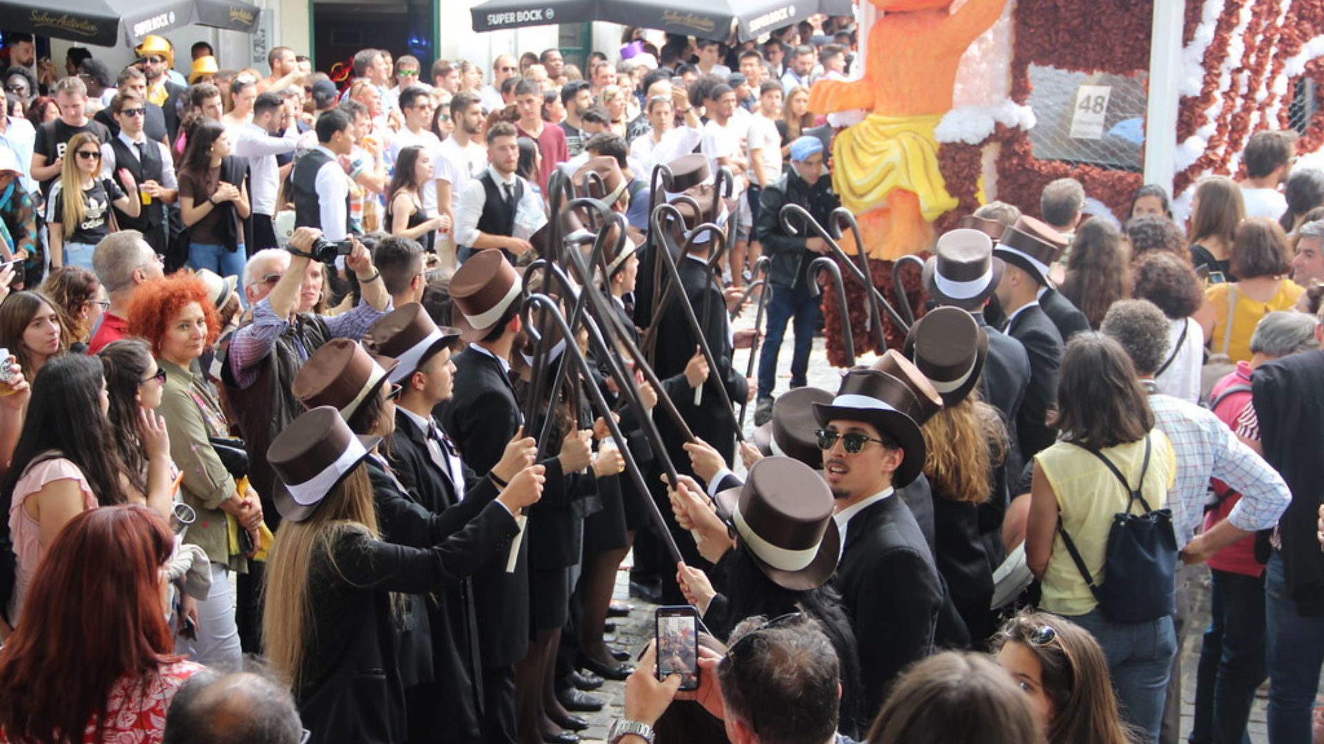A large crowd of university students in black academic attire and some wearing hats, gathered outdoors during a daytime parade, with a large, elaborate red float topped with a figure in the background.