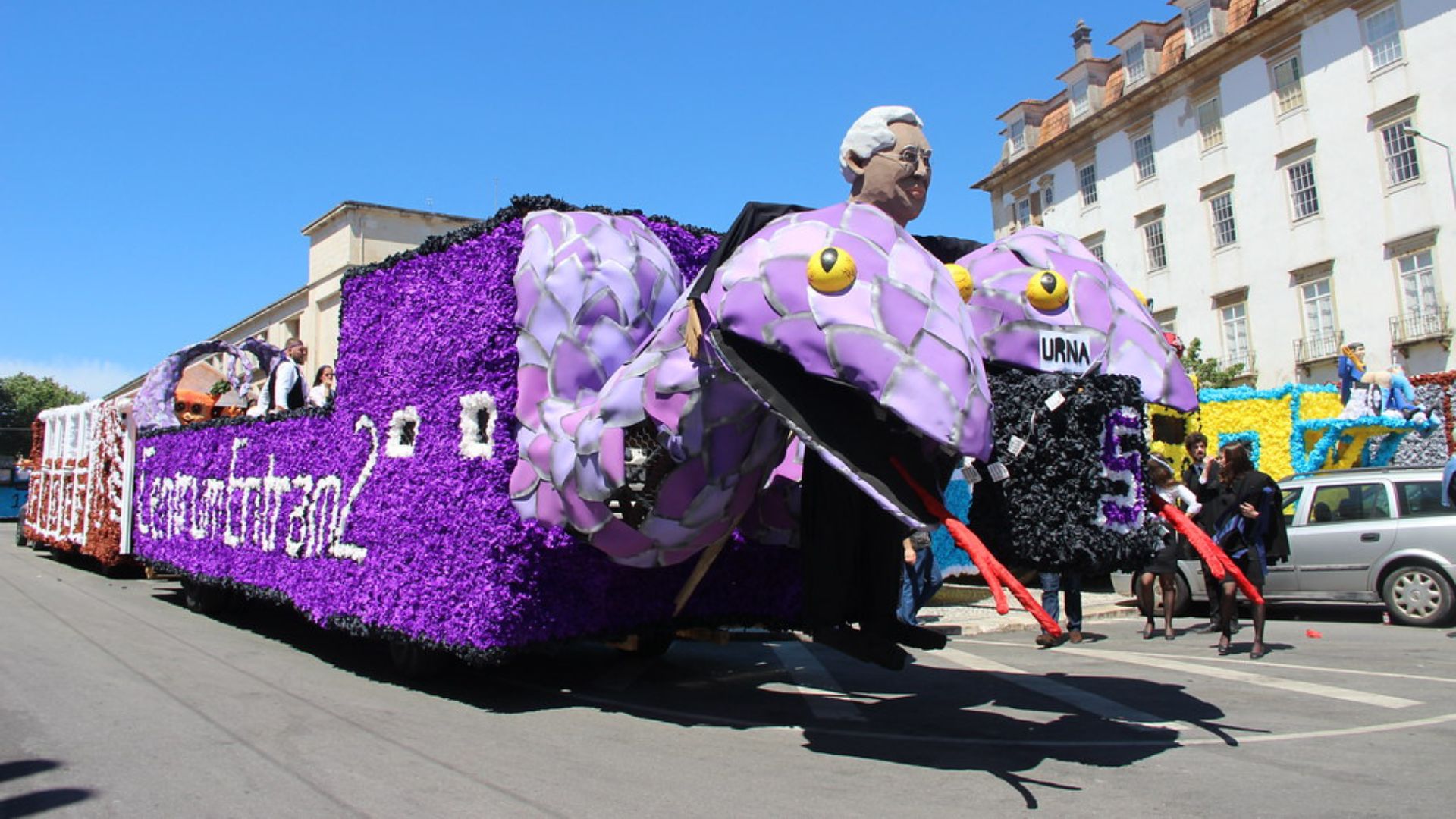 A vibrant purple float shaped like a large serpent with a human bust on its head, featuring prominent eyes and scales, parades down a sunny street during the Cortejo da Queima das Fitas in Coimbra, Portugal. The float is adorned with purple flowers and text, and spectators line the street, with historic buildings visible in the background.