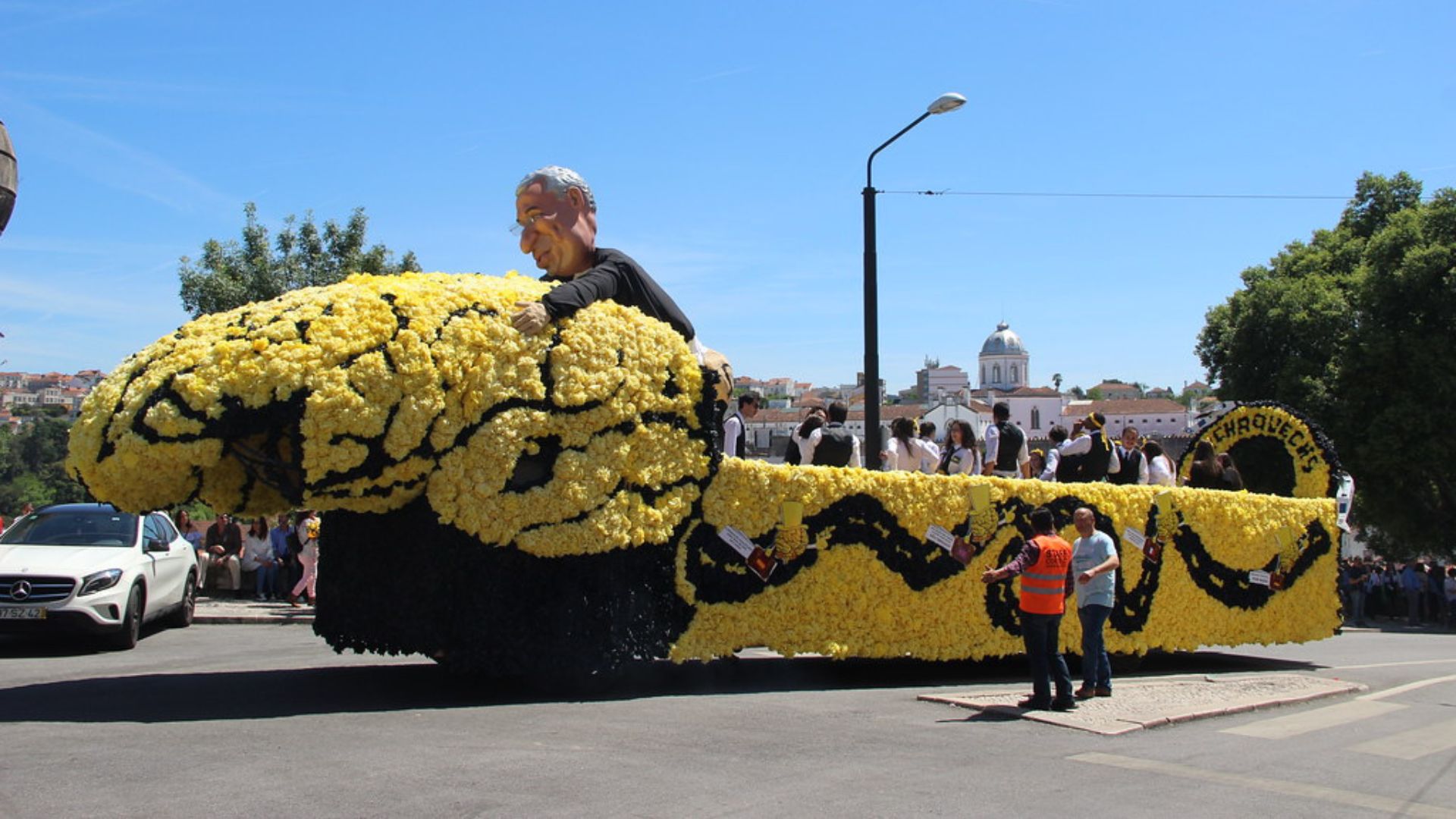 A large, elaborate parade float shaped like a yellow brain with a man's figure emerging from it, decorated with black accents, moves down a street under a clear blue sky. Students in traditional dark academic attire are visible on and around the float, with historic buildings and trees in the background.