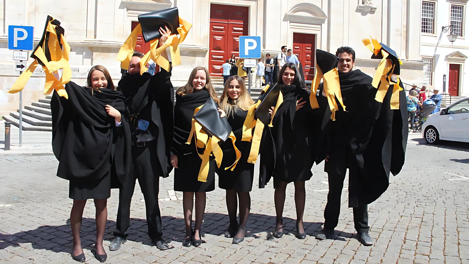 A group of smiling students in black academic robes and suits with yellow ribbon, holding folders, standing in front of a building during the Queima das Fitas festival in Coimbra, Portugal.