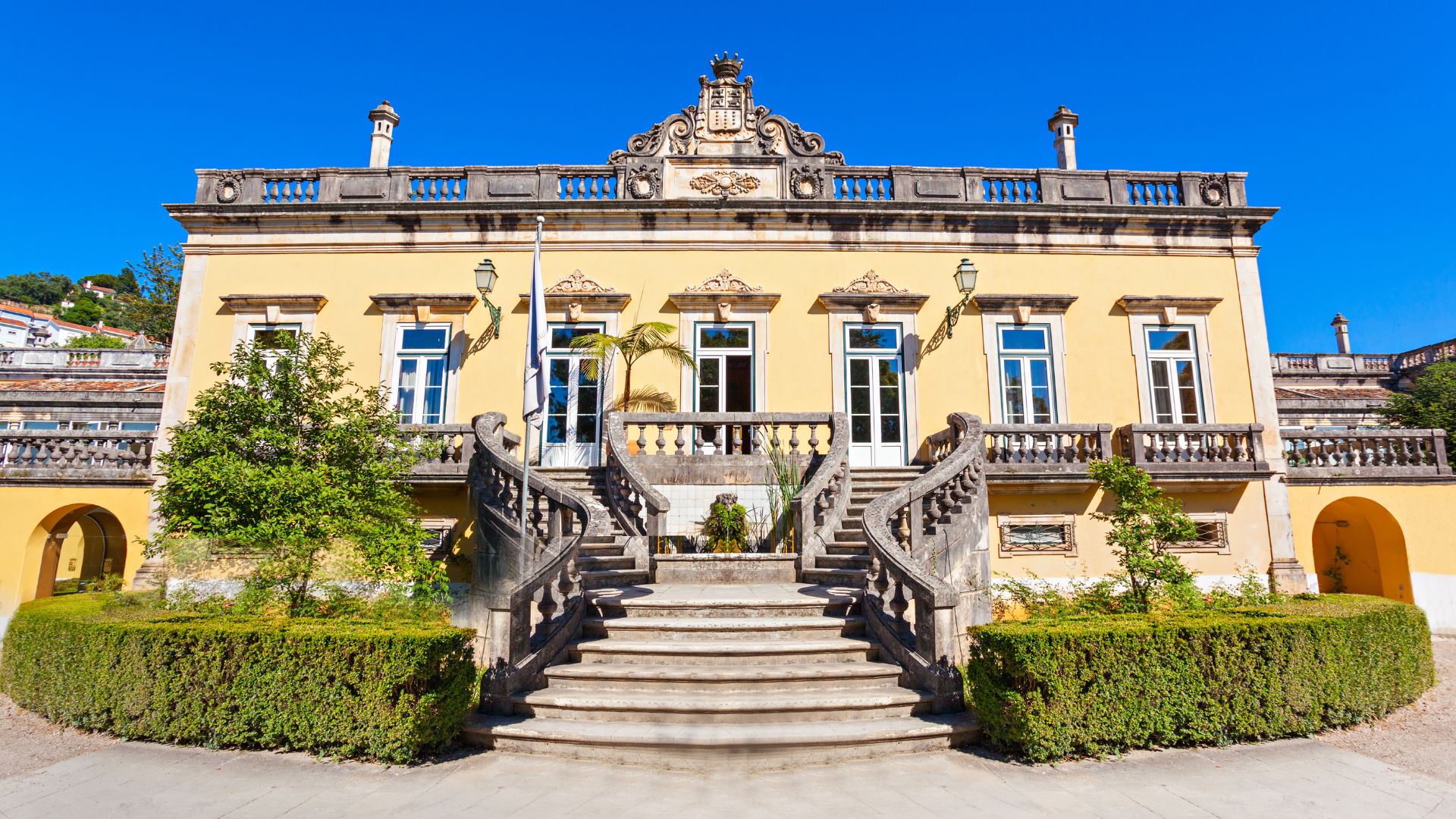 A grand yellow palace with intricate architectural details and a prominent set of double-curved stone staircases leading up to its main entrance, surrounded by manicured hedges and lush greenery under a clear blue sky.