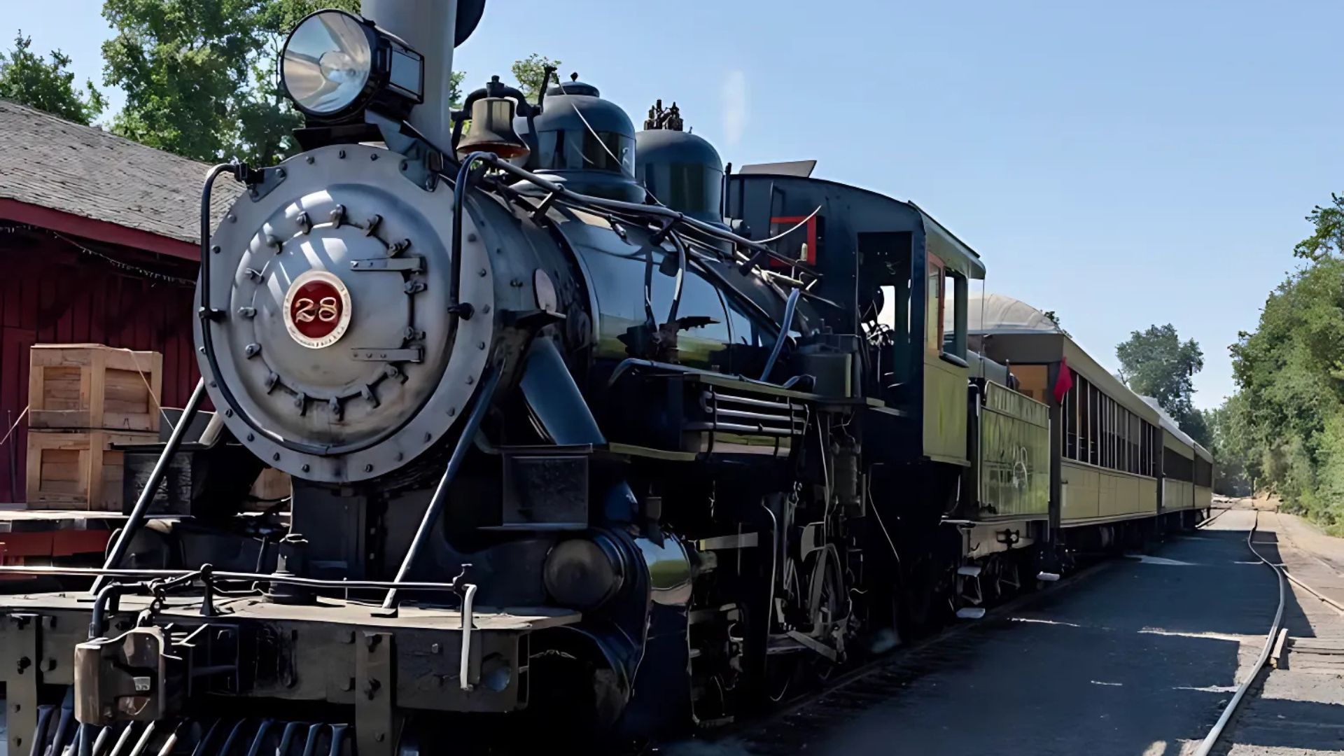 A historic black steam locomotive, Sierra Railway No. 28, with the number "28" visible on its front, is prominently featured on a track at Railtown 1897 State Historic Park. The train is coupled with passenger cars extending into the background on the right, while a red building and wooden crates are visible on the left. The scene is set outdoors under a clear sky.