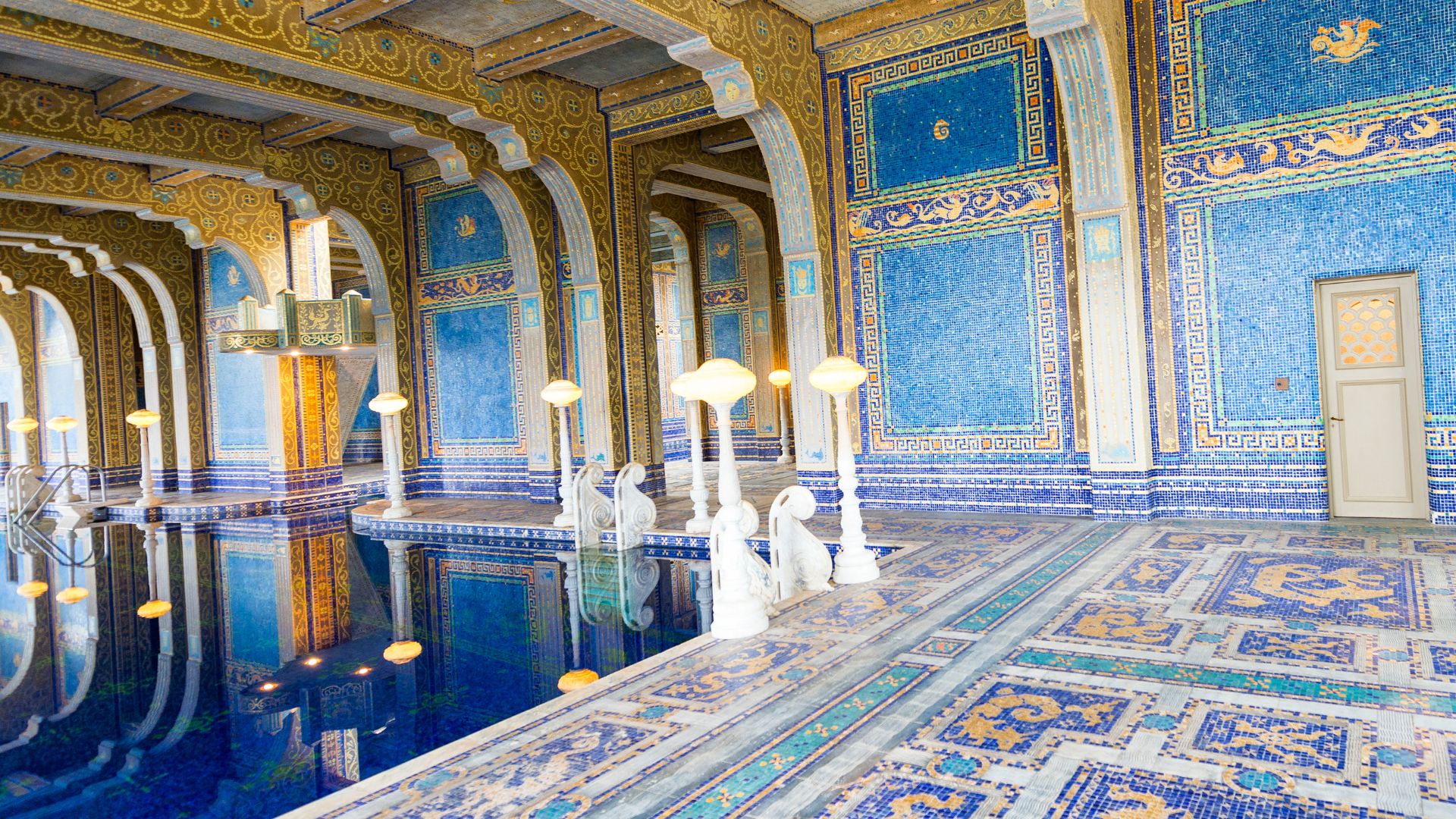 The image shows the opulent indoor Roman Pool at Hearst Castle, featuring intricate blue and gold mosaic tiles, classical statues, and ornate arches, reflecting the grandeur of ancient Roman baths. 