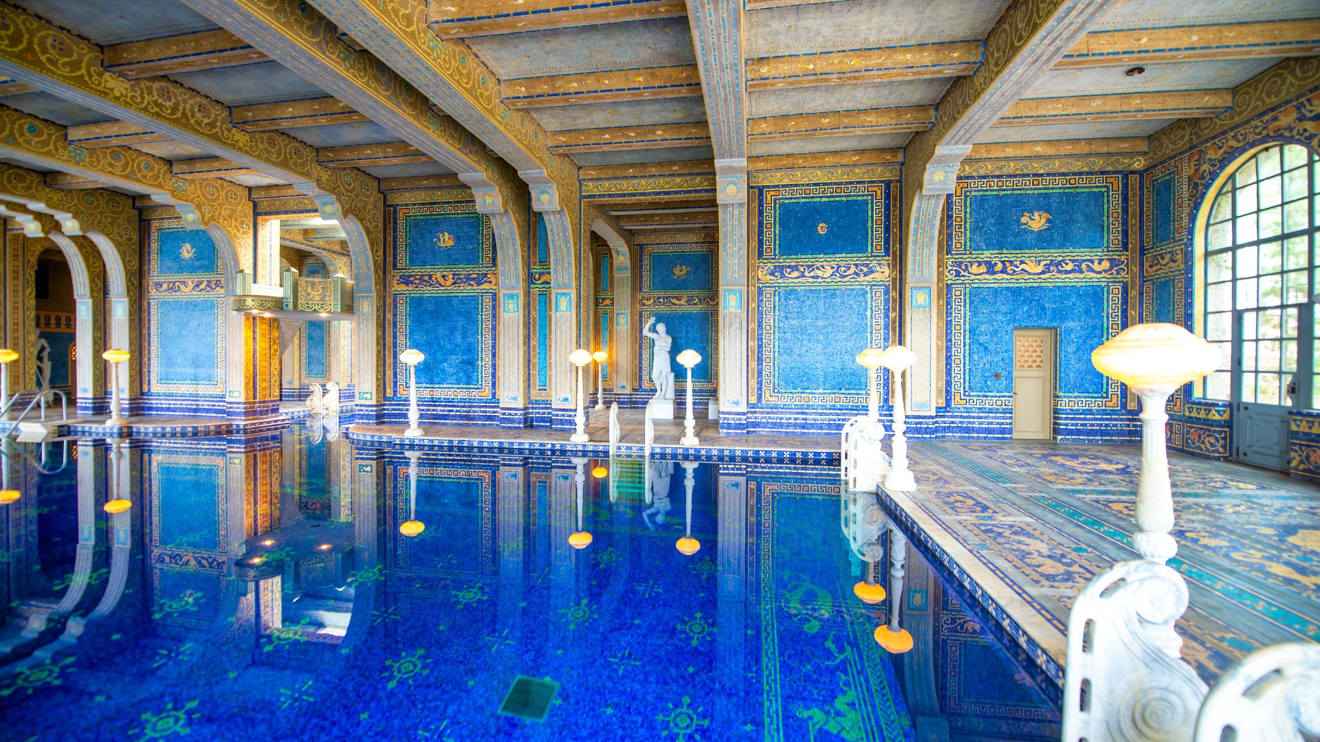 A lavish indoor swimming pool, known as the Roman Pool at Hearst Castle, featuring vibrant blue and gold mosaic tiles, illuminated by soft lighting from ornate lamps and natural light from a large arched window. The pool is surrounded by classical statues and grand architectural details, reflecting in the deep blue water. 