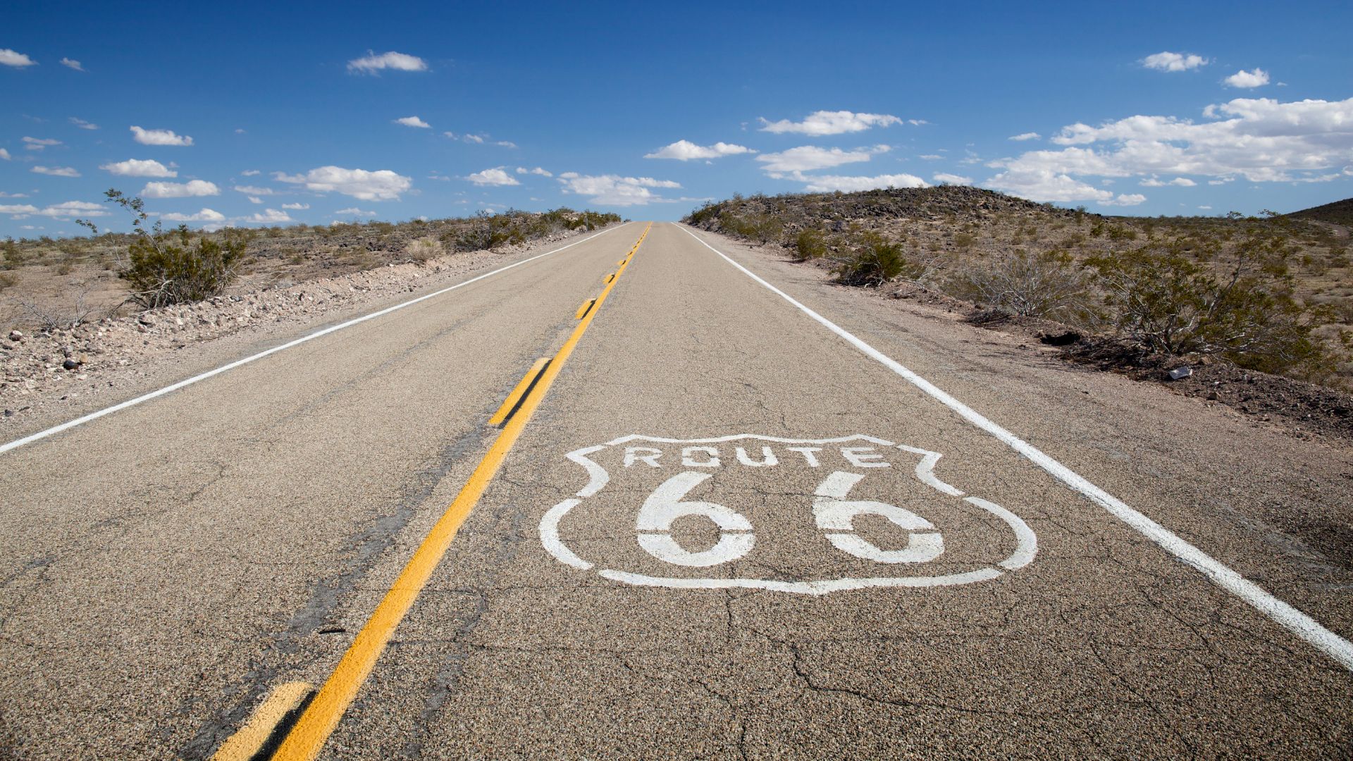 A low-angle shot of a long, straight stretch of empty asphalt road, with the iconic white "ROUTE 66" shield painted prominently on the surface in the foreground. The road extends into the distance under a clear blue sky with scattered white clouds, flanked by dry, brush-covered hills on either side.