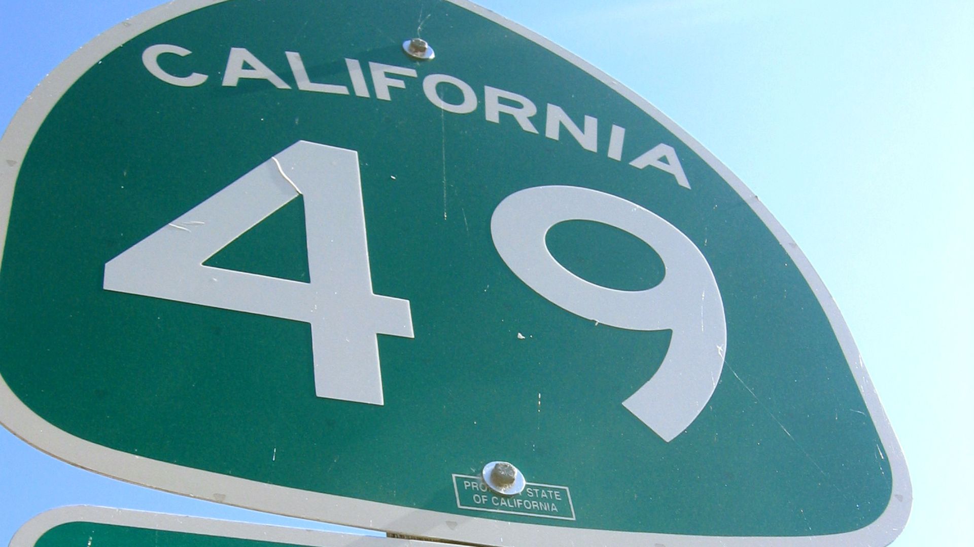 A green, shield-shaped California State Route 49 highway sign with "CALIFORNIA" written above the large white numbers "49."