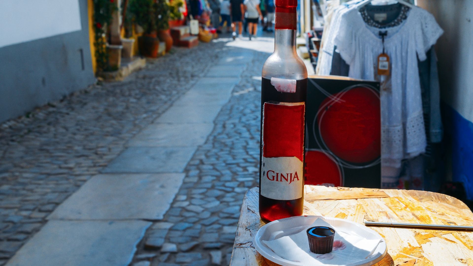 A bottle of Ginja de Obidos and a small chocolate cup on a table, with a cobbled street and buildings in the background.