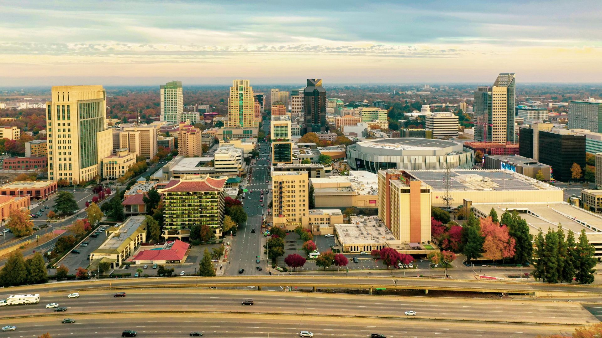 An aerial view of a bustling city skyline at dusk, showcasing numerous tall buildings, a prominent bridge, and a wide roadway with vehicles, all under a colorful sky with hints of clouds. The city is surrounded by trees and appears to be a major urban center.